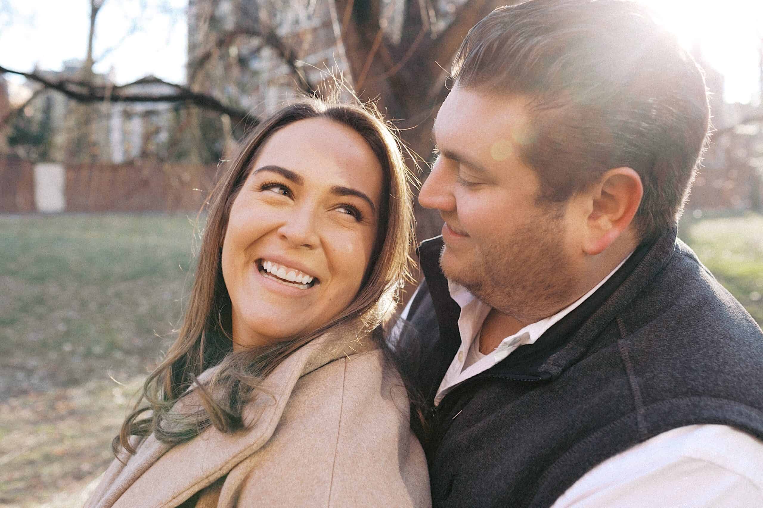 A woman smiles while a man sits close behind her, looking at her. They are outdoors with sunlight in the background.