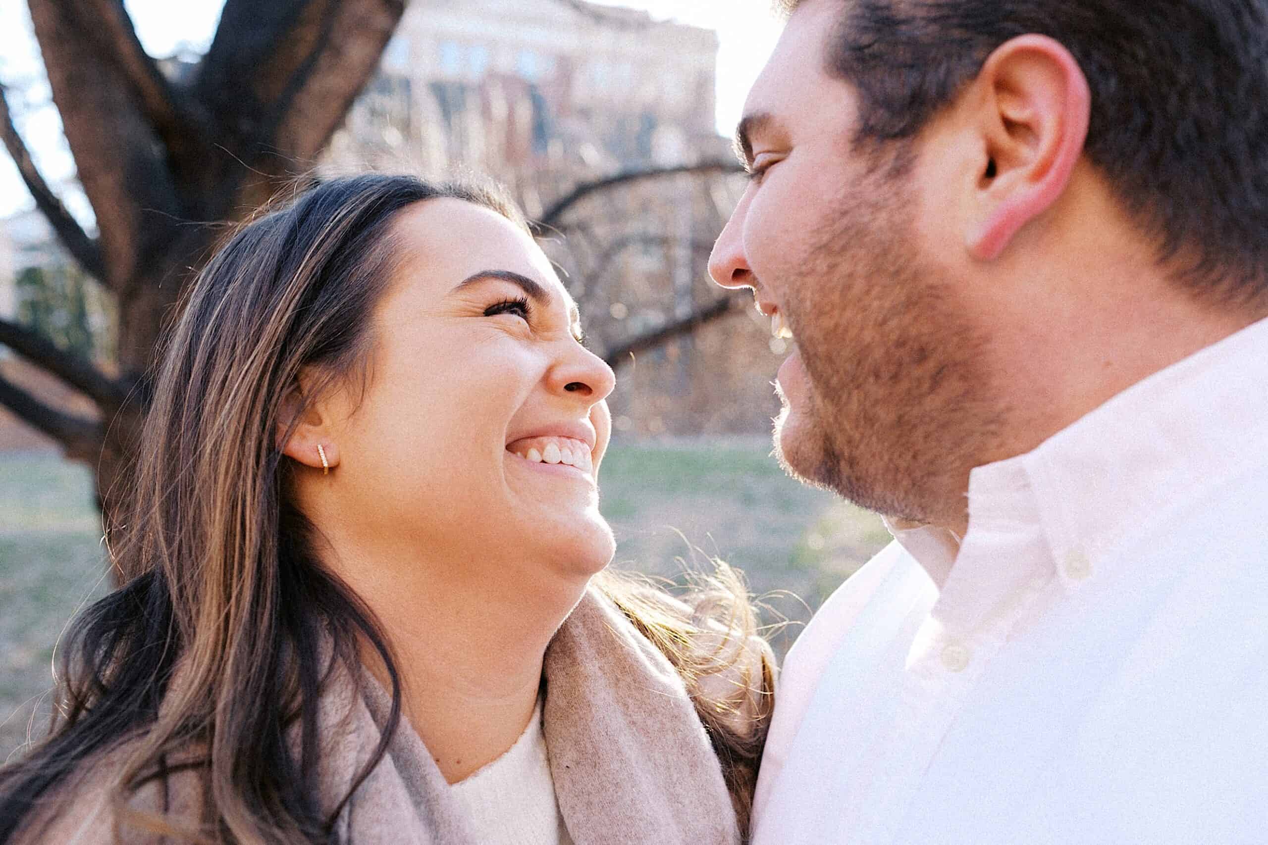 A woman and man standing outdoors, facing each other and smiling, with trees and a building visible in the background.