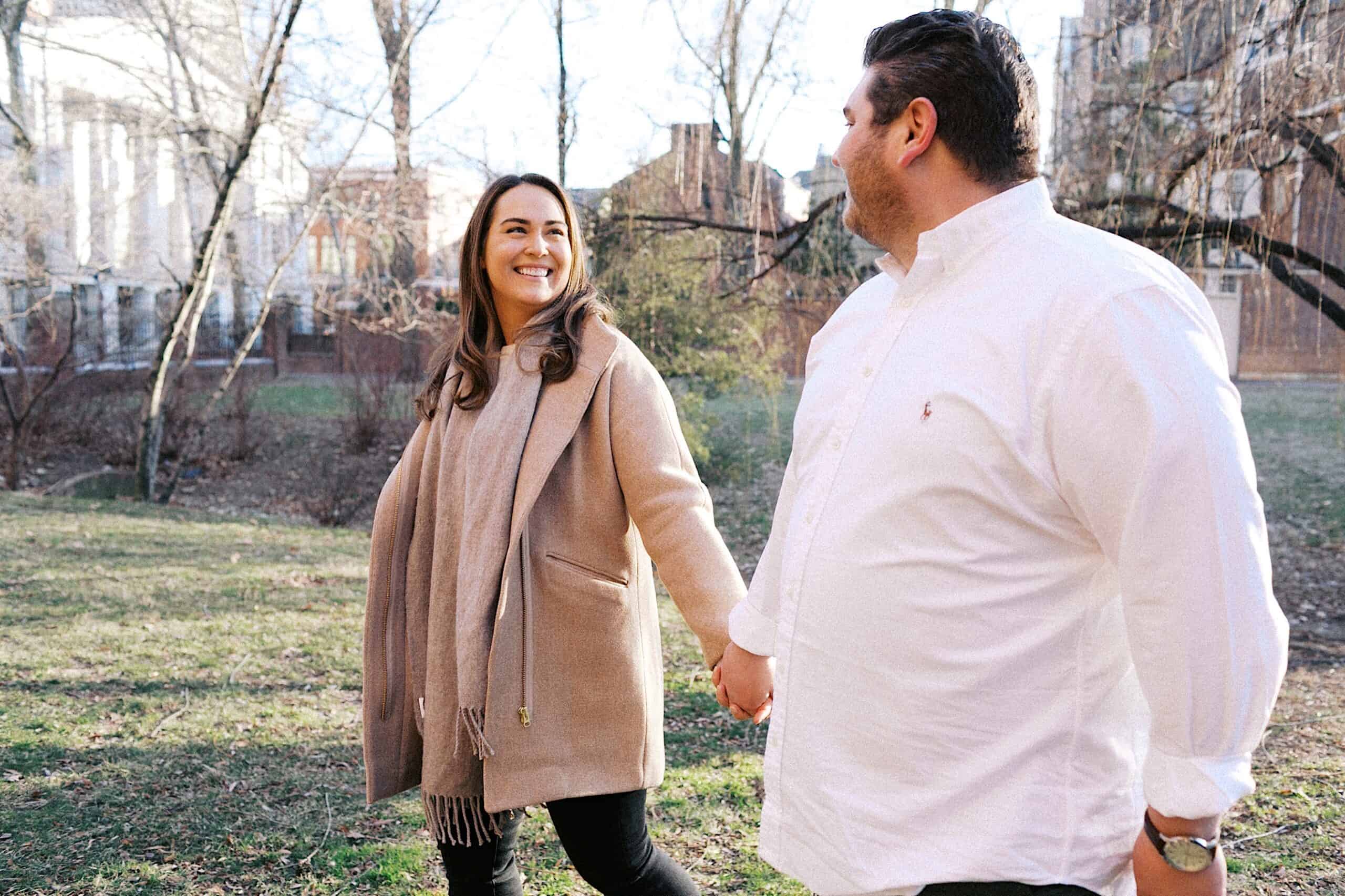 A woman and man hold hands and walk outdoors in a park on a sunny day, both dressed in casual winter clothing and smiling at each other.
