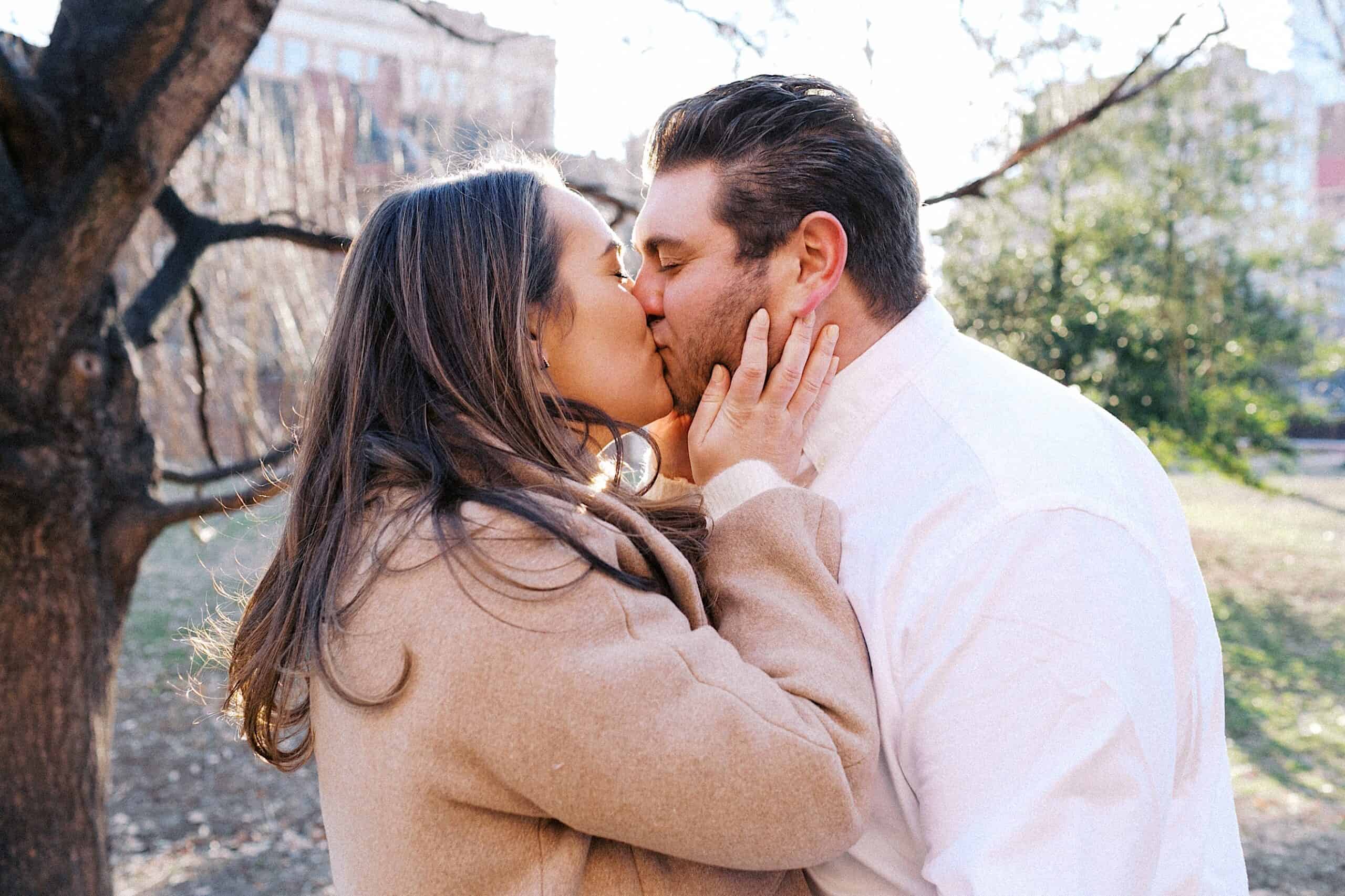 A couple stands outdoors in a park, wearing winter coats, as they kiss each other on the lips.
