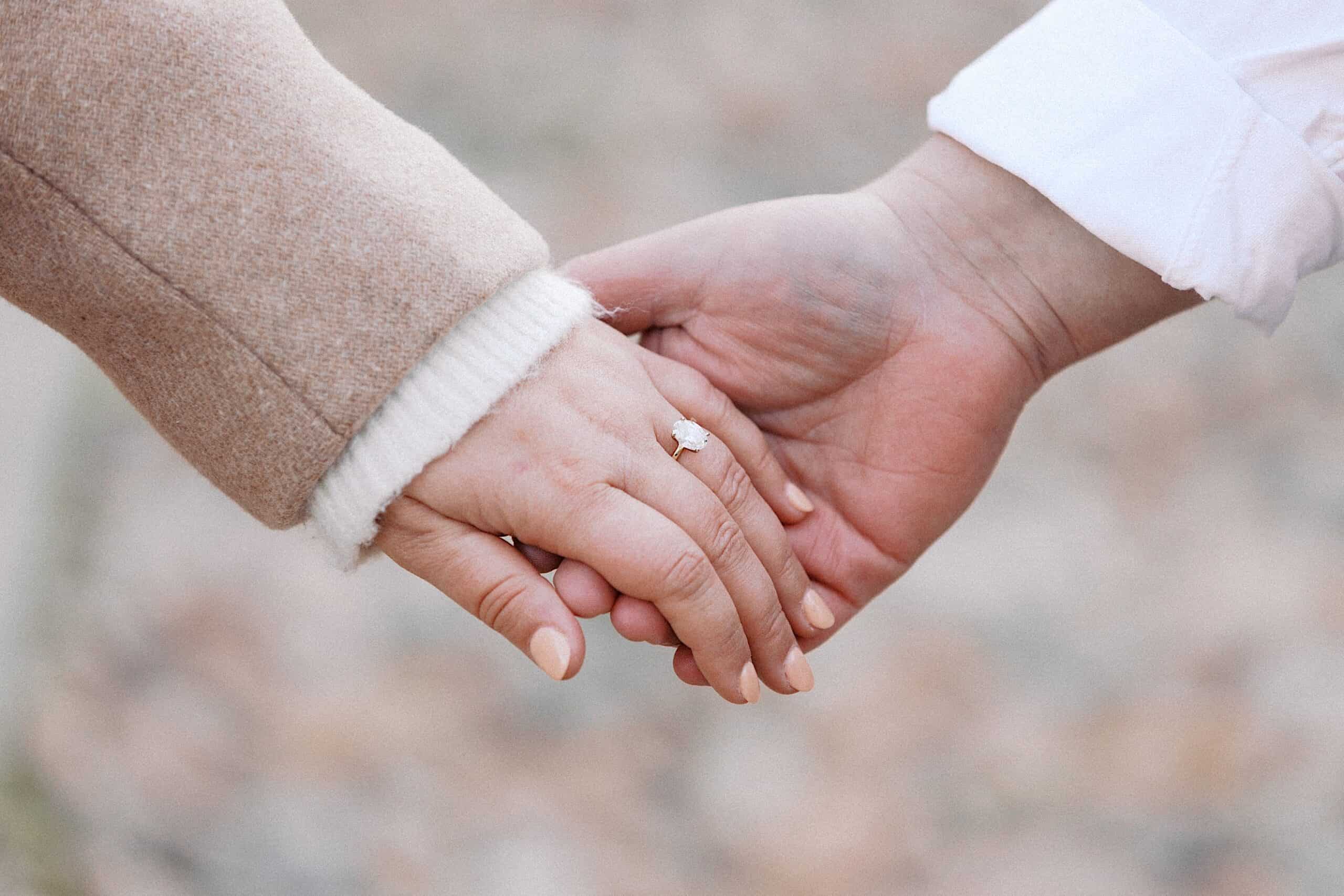 Close-up of two people holding hands, one wearing a beige coat and an engagement ring, the other in a white sleeve.