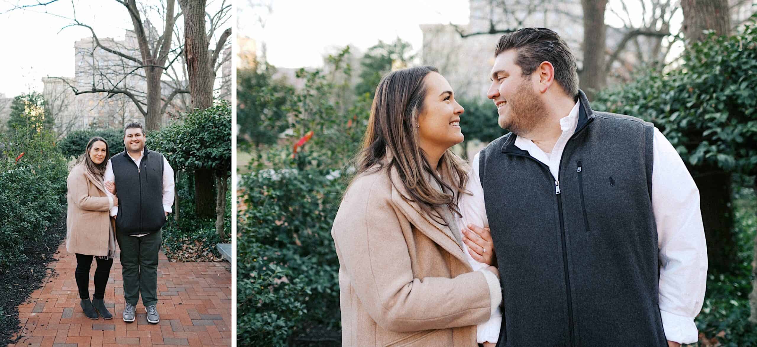 A couple stands closely together on a brick path in a park, smiling at each other with greenery and trees in the background.