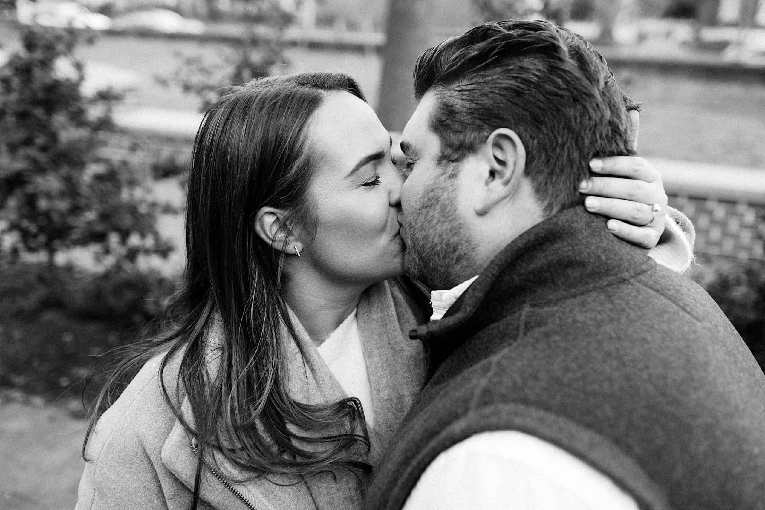 A couple is kissing outdoors, with the woman holding the man's head close. The image is black and white.