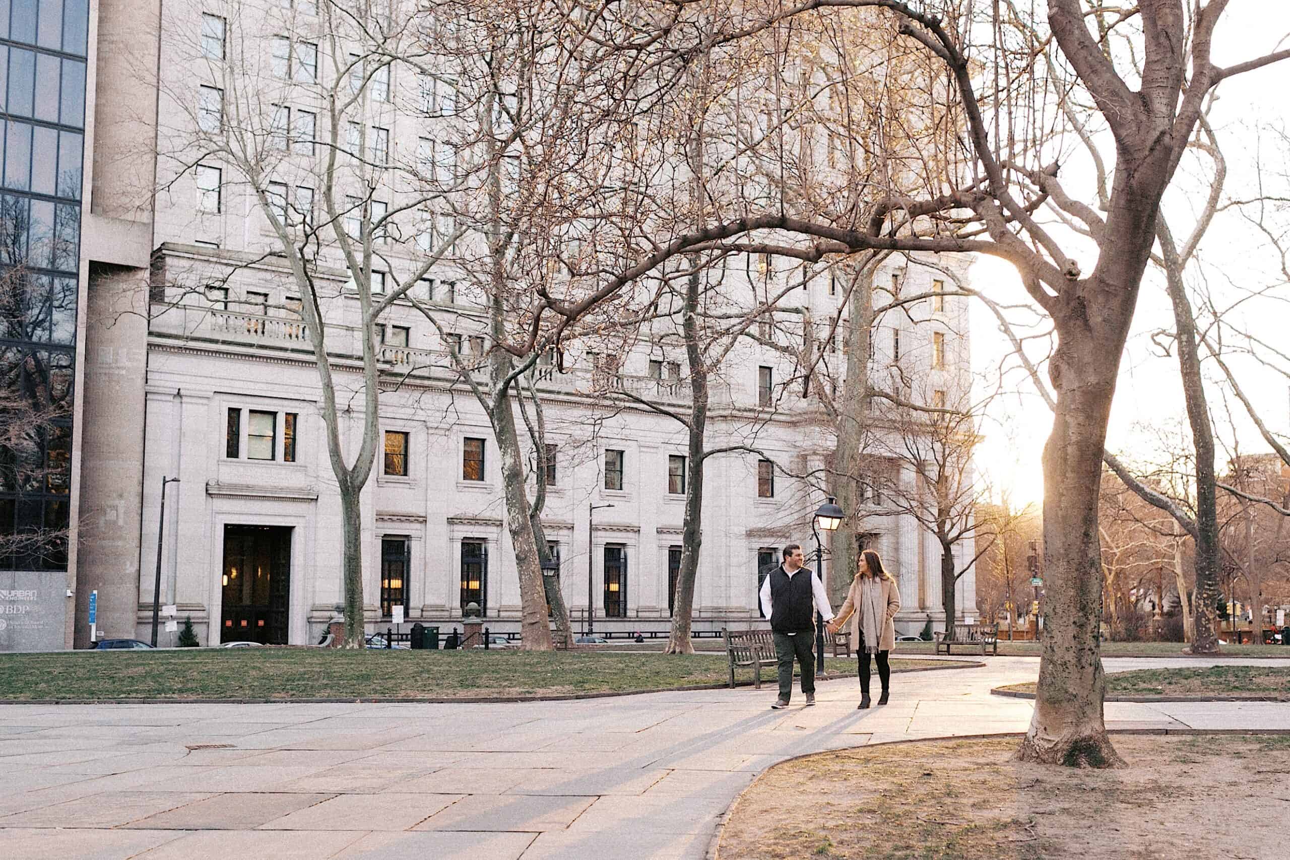 Two people walk together on a paved path in a city park with bare trees and a large stone building in the background at sunset.