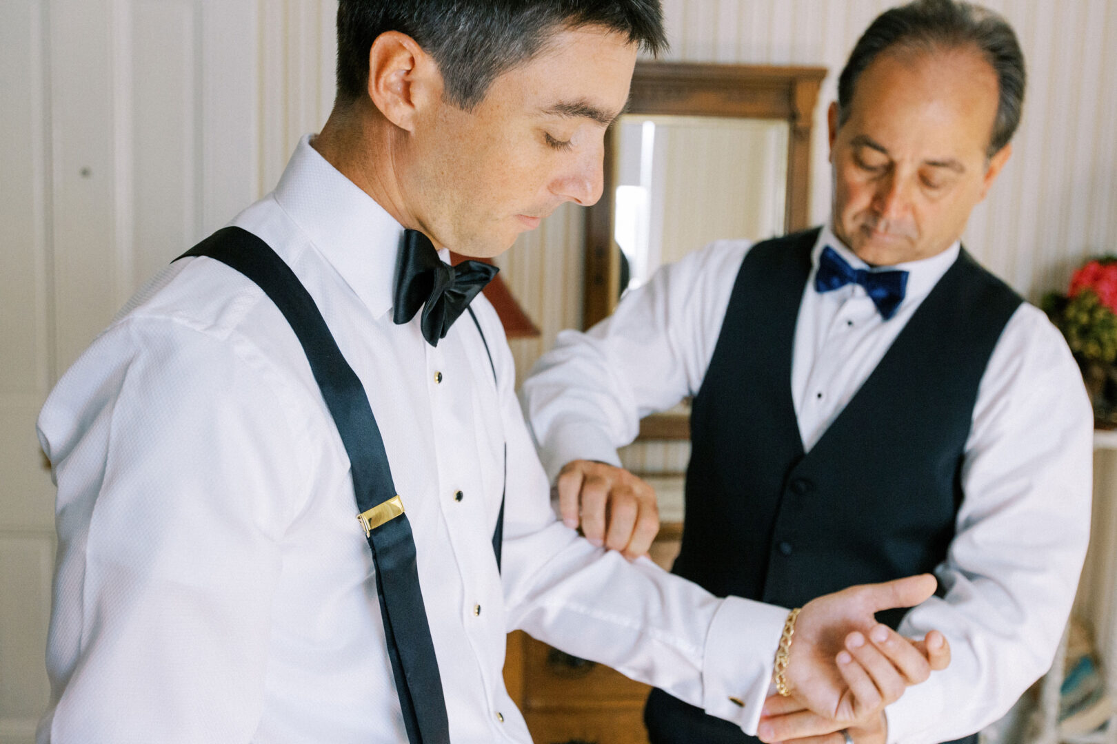 Two men in dress shirts and bow ties, one looking down adjusting sleeves, the other assisting, both wearing black suspenders. A mirror and furniture are in the background.