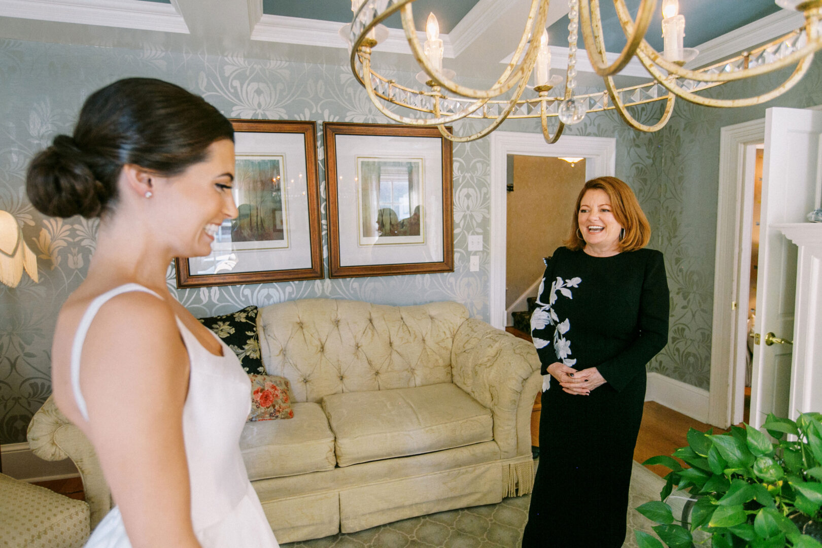 A bride in a white dress smiles at a woman in a black dress, surrounded by patterned wallpaper, framed pictures, and a chandelier. Their laughter resonates with the elegance of a Cape May Convention Hall wedding.