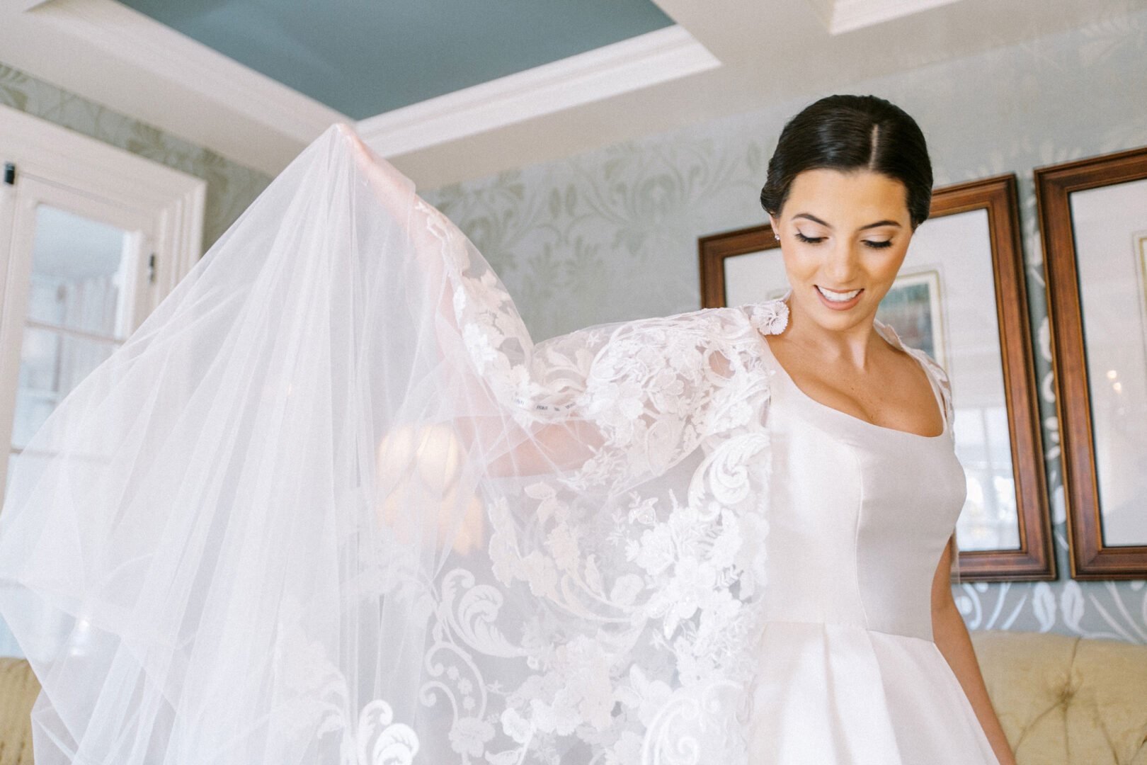 Bride in a white dress holding her veil, standing in a room with patterned wallpaper and framed pictures, recalling memories of her enchanting Cape May Convention Hall wedding.