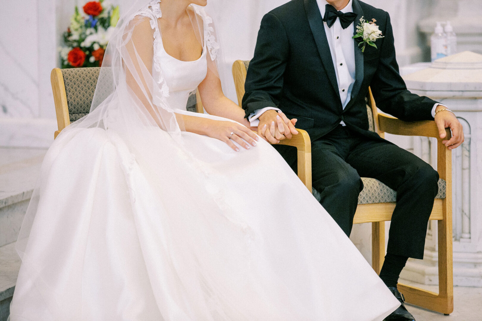 The bride, in her stunning white gown and veil, holds hands with the groom, dashing in his black tuxedo. They are seated on upholstered chairs in a formal setting at their Cape May Convention Hall wedding.