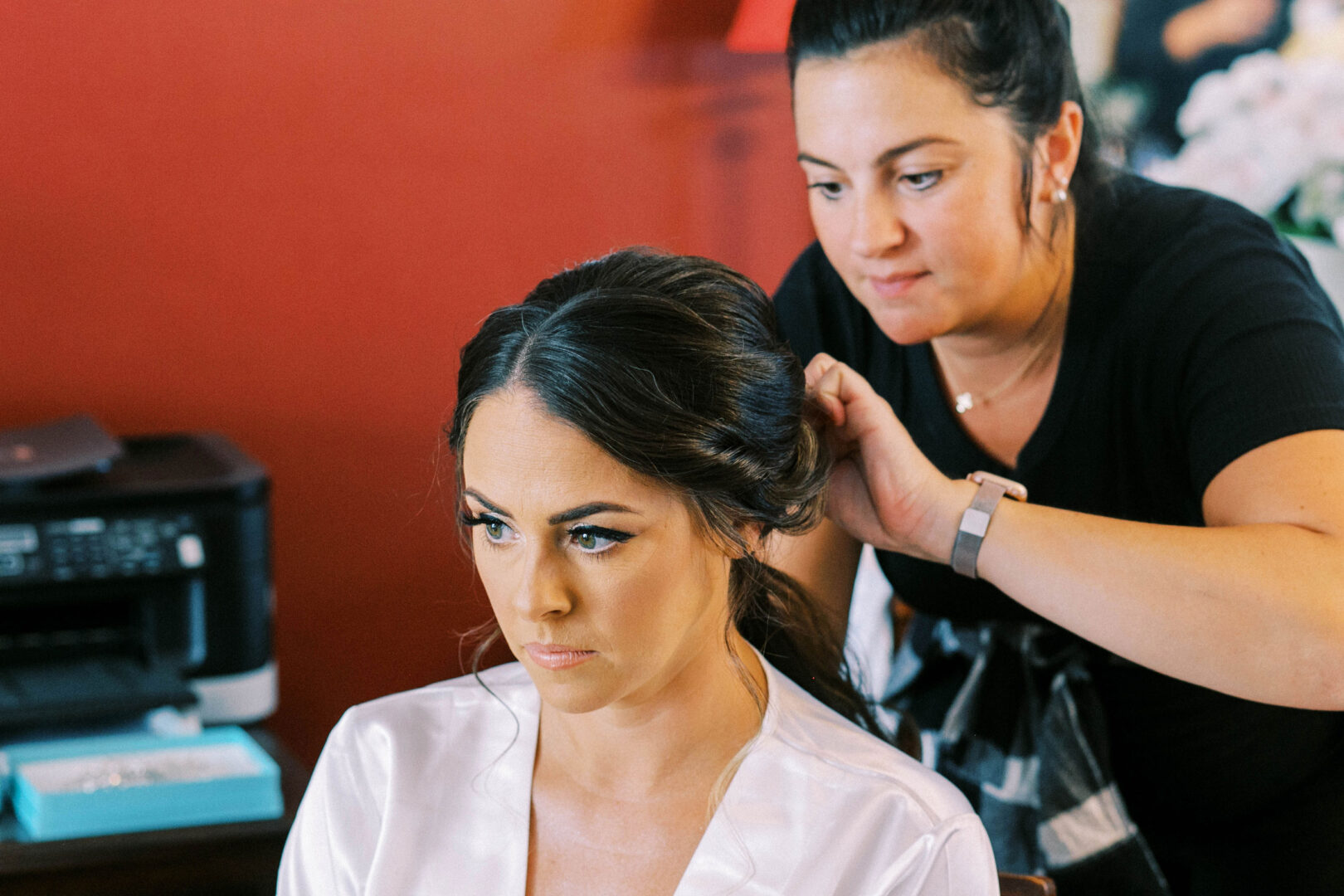 In a room with red walls, the hairstylist meticulously arranges the woman's hair for her wedding photos. Seated in a white robe, she radiates anticipation at the Mansion on Main Street.