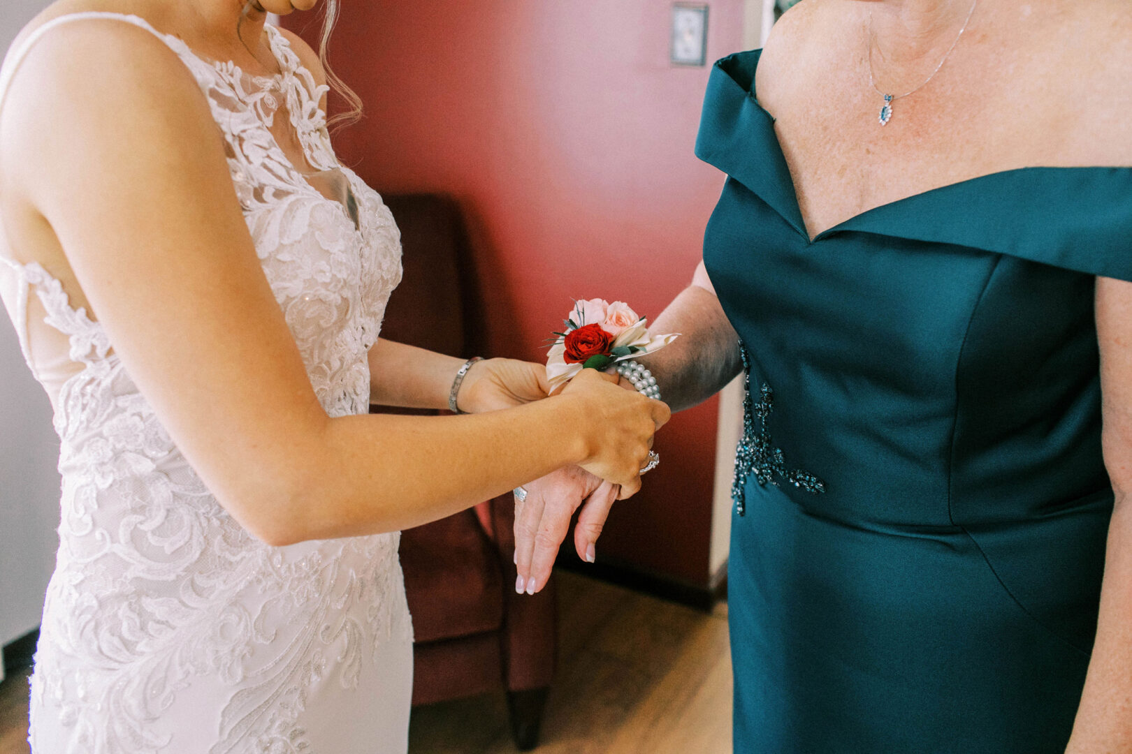 In the Mansion on Main Street, a bride in a white lace dress gently places a corsage on the wrist of a woman in a dark teal gown, capturing a timeless moment for their wedding photos against the backdrop of rich red walls.