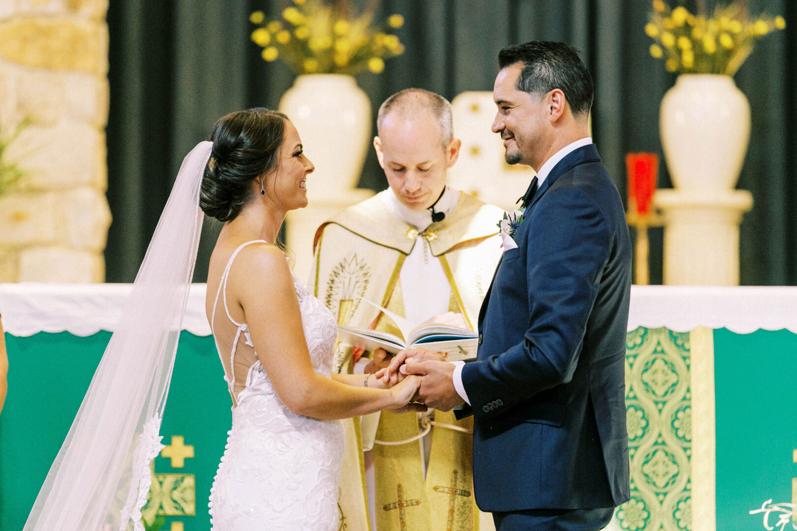 A bride and groom stand facing each other, smiling, while a priest reads from a book during their wedding ceremony at the Mansion on Main Street. They are surrounded by elegant decor, capturing the perfect moment in their cherished wedding photos.