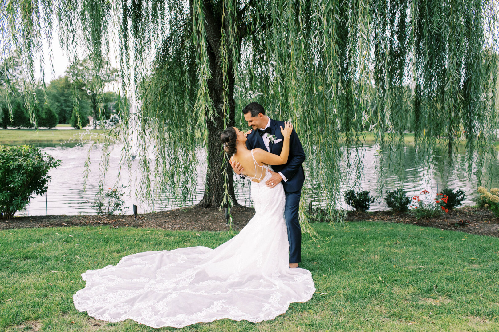 The bride and groom elegantly pose under a willow tree by a serene lake at the Mansion on Main Street, with her in a stunning white dress and him in a sharp dark suit, their wedding photos capturing the beauty of nature and love.
