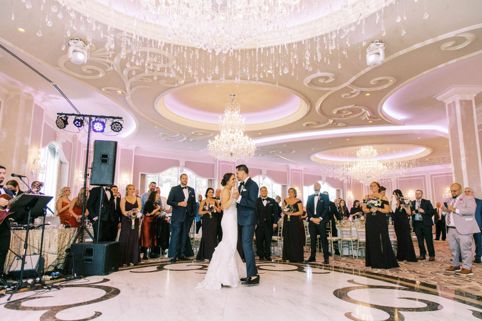 At the Mansion on Main Street, a couple dances gracefully at their wedding reception beneath sparkling chandeliers, as guests capture timeless wedding photos.