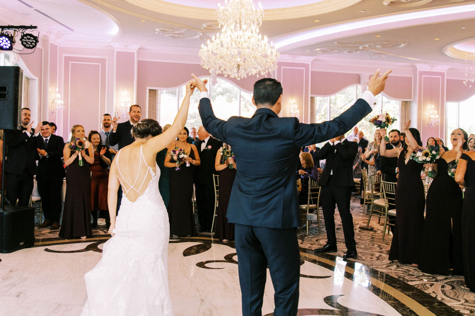 A newlywed couple raises their arms while facing applauding guests in the elegant ballroom of the Mansion on Main Street, where a large chandelier adds a touch of romance to their unforgettable wedding celebration.