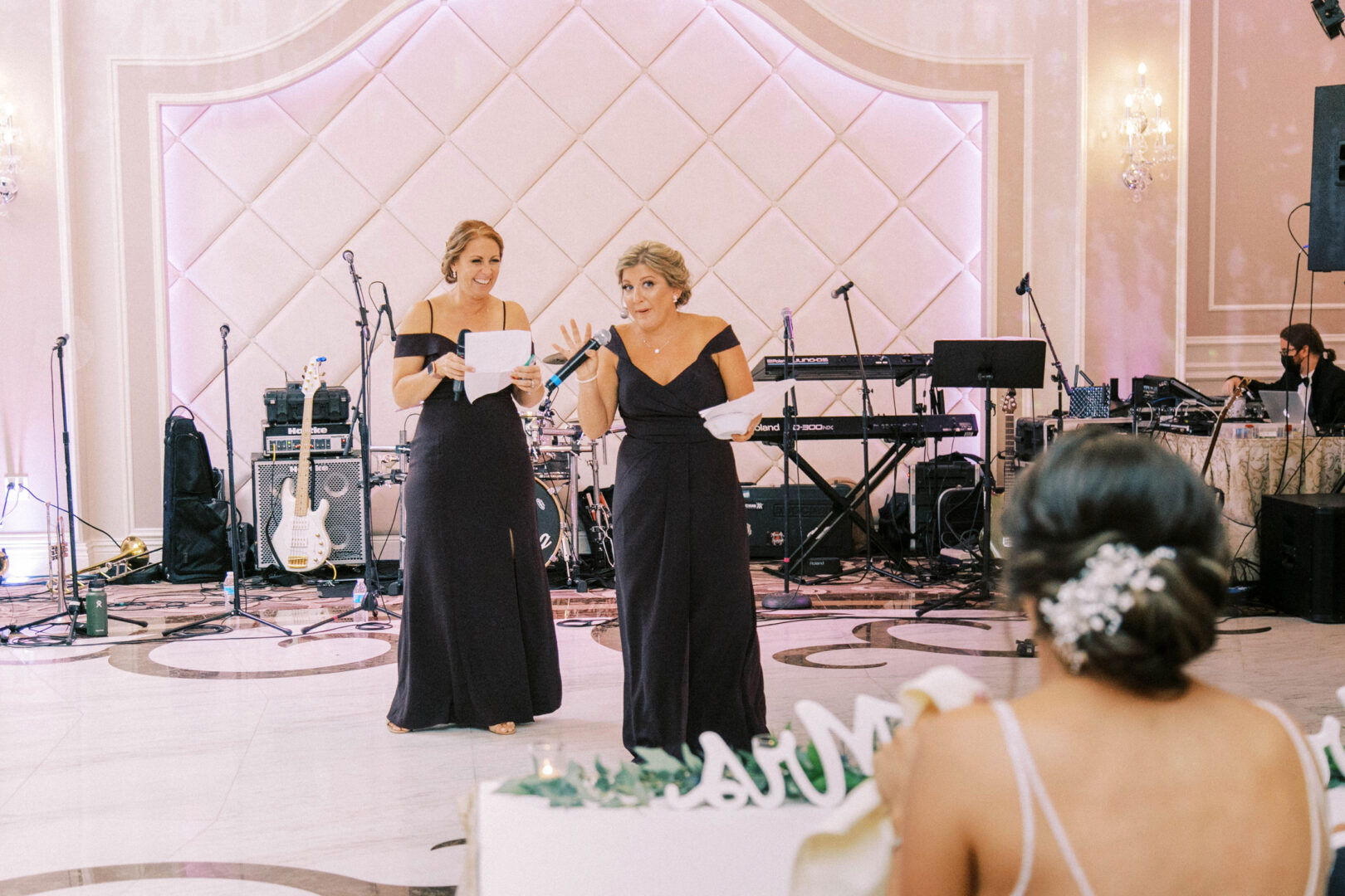 Two women in formal dresses speak into microphones at an elegant indoor wedding, with musical equipment in the background. The Mansion on Main Street provides a stunning backdrop, perfect for capturing unforgettable wedding photos.