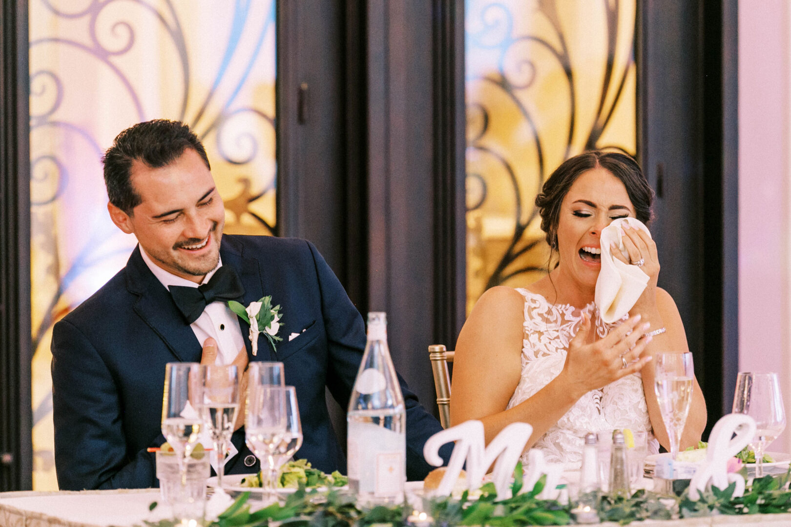 At their wedding at the Mansion on Main Street, a bride and groom sit at a table, laughing. The bride wipes tears with a napkin as glasses and a bottle glimmer nearby, capturing perfect moments for their wedding photos.