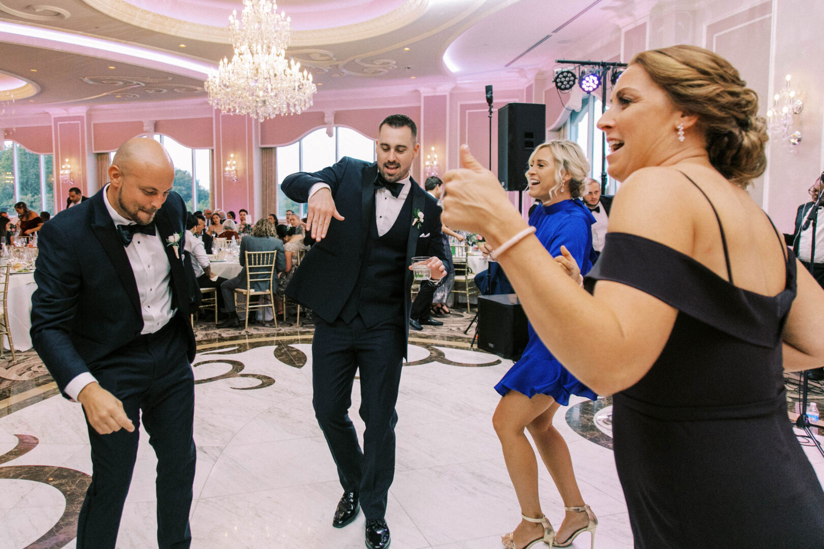 Four people in formal attire are dancing energetically on the ballroom floor, a dazzling chandelier overhead, capturing the joyous essence of a wedding at the Mansion on Main Street.