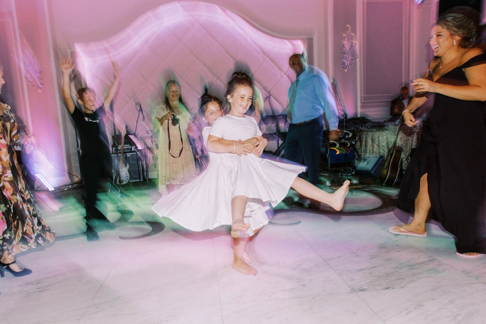 Two children are dancing joyfully in a lively room, surrounded by people on a gleaming marble floor at the Mansion on Main Street. It's as if they're creating their own magical moment amid the unfolding wedding celebration captured in treasured photos.