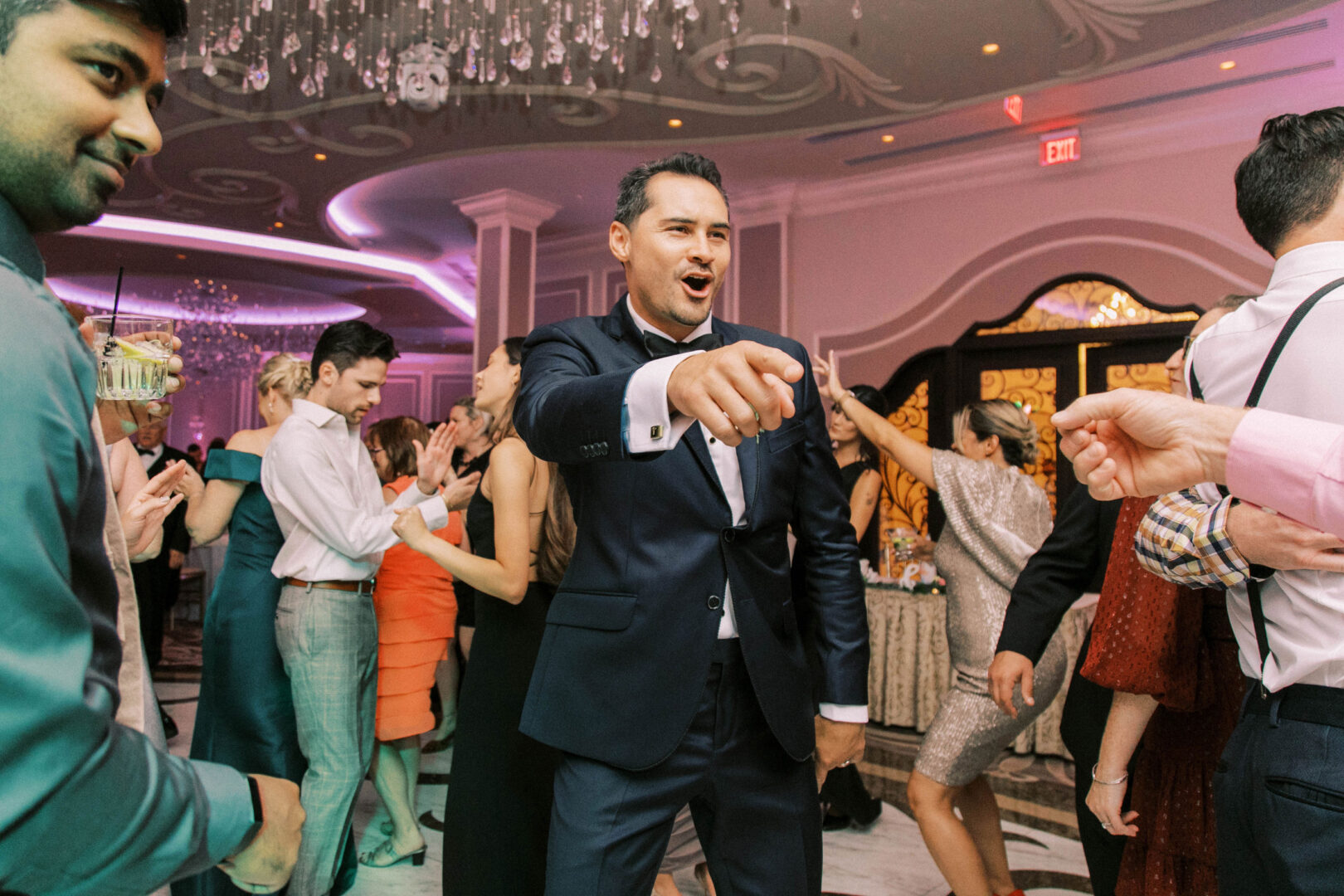 At the Mansion on Main Street, a man in a suit energetically points and dances at an elegant wedding, surrounded by guests clapping and dancing in a lavishly decorated ballroom captured beautifully in wedding photos.