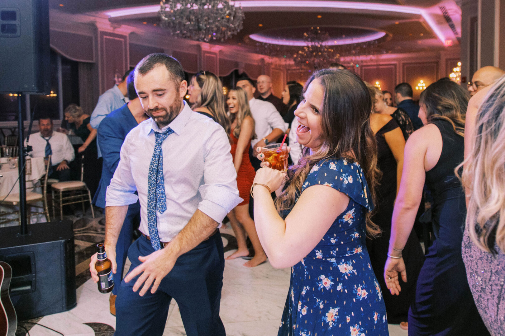 Guests are dancing and holding drinks at a lively indoor wedding party with dim lighting and elegant decor, capturing perfect wedding photos.