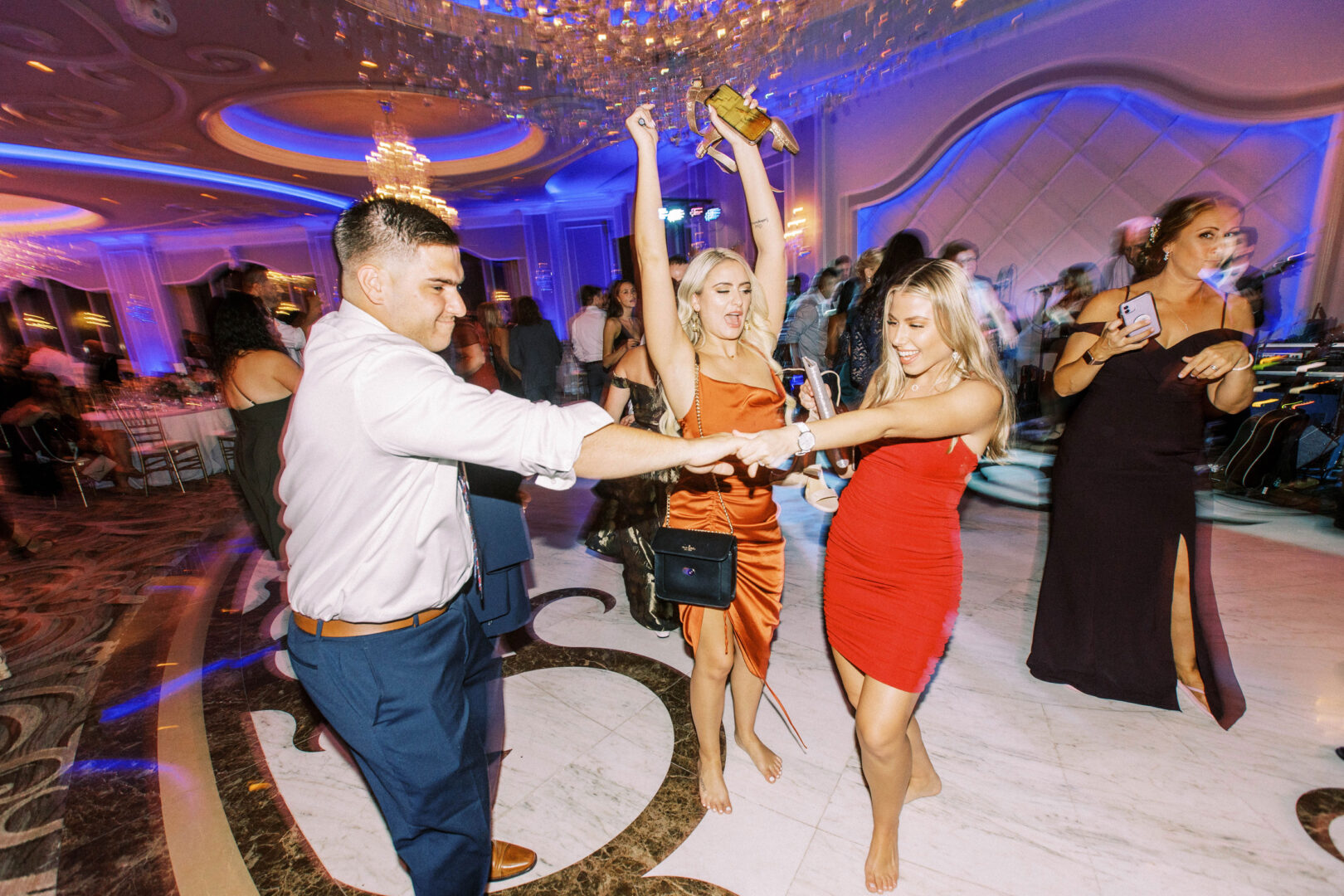 In the elegant ballroom of the Mansion on Main Street, where chandeliers cast a warm glow, people dance energetically. Two women in red dresses take center stage, creating picture-perfect wedding photos as they are surrounded by lively guests.
