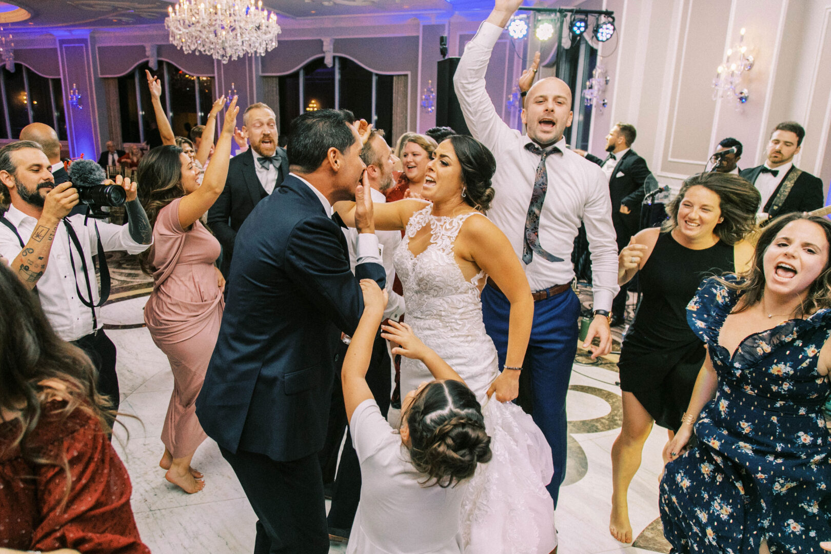 A joyful crowd, including a bride in a white dress and several people wearing formal attire, are dancing energetically on the ballroom floor at the Mansion on Main Street, capturing perfect wedding photos.