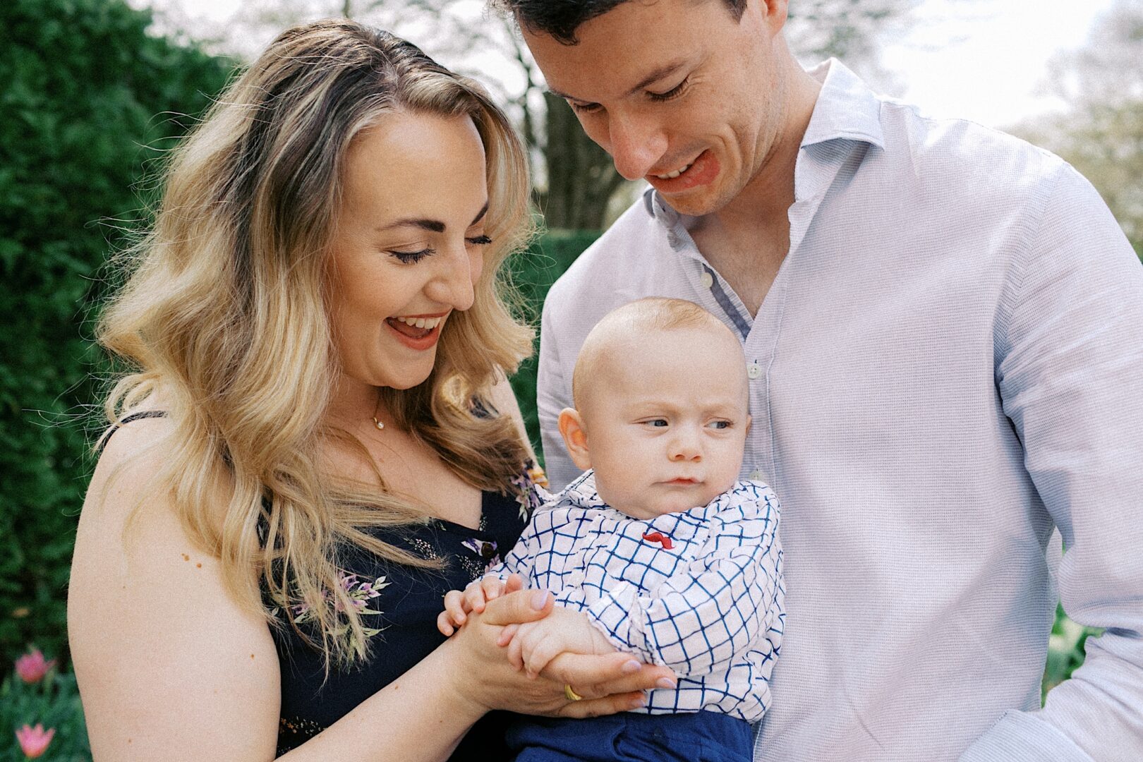 A woman and man are all smiles as they bask in the perfect spring day, holding their baby amidst lush greenery—capturing ideal family photos outdoors.