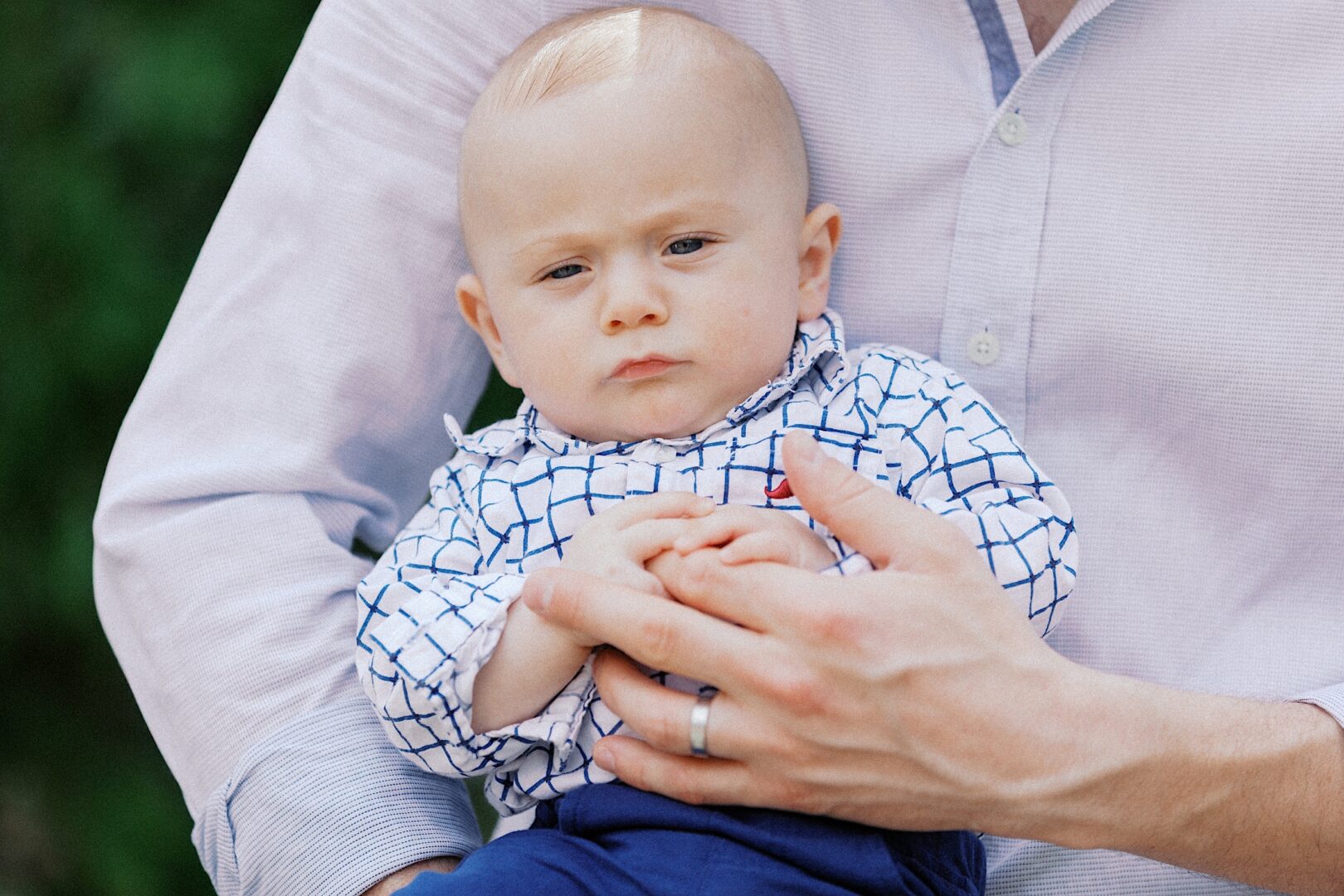 A baby in a patterned shirt is lovingly cradled by an adult in a light button-up, their ring glinting softly. With the sun-dappled greenery as a backdrop, it captures the essence of perfect spring family photos.