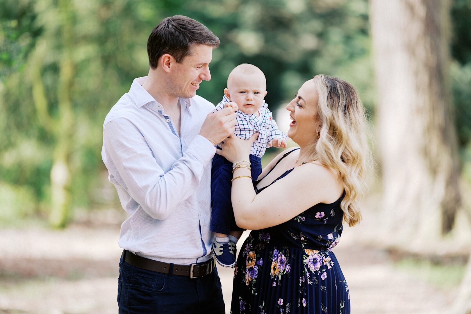 A man and woman hold their baby outdoors, basking in the joy of perfect spring family photos. The man smiles warmly as he touches the baby's hand, while the woman gazes lovingly at their child.