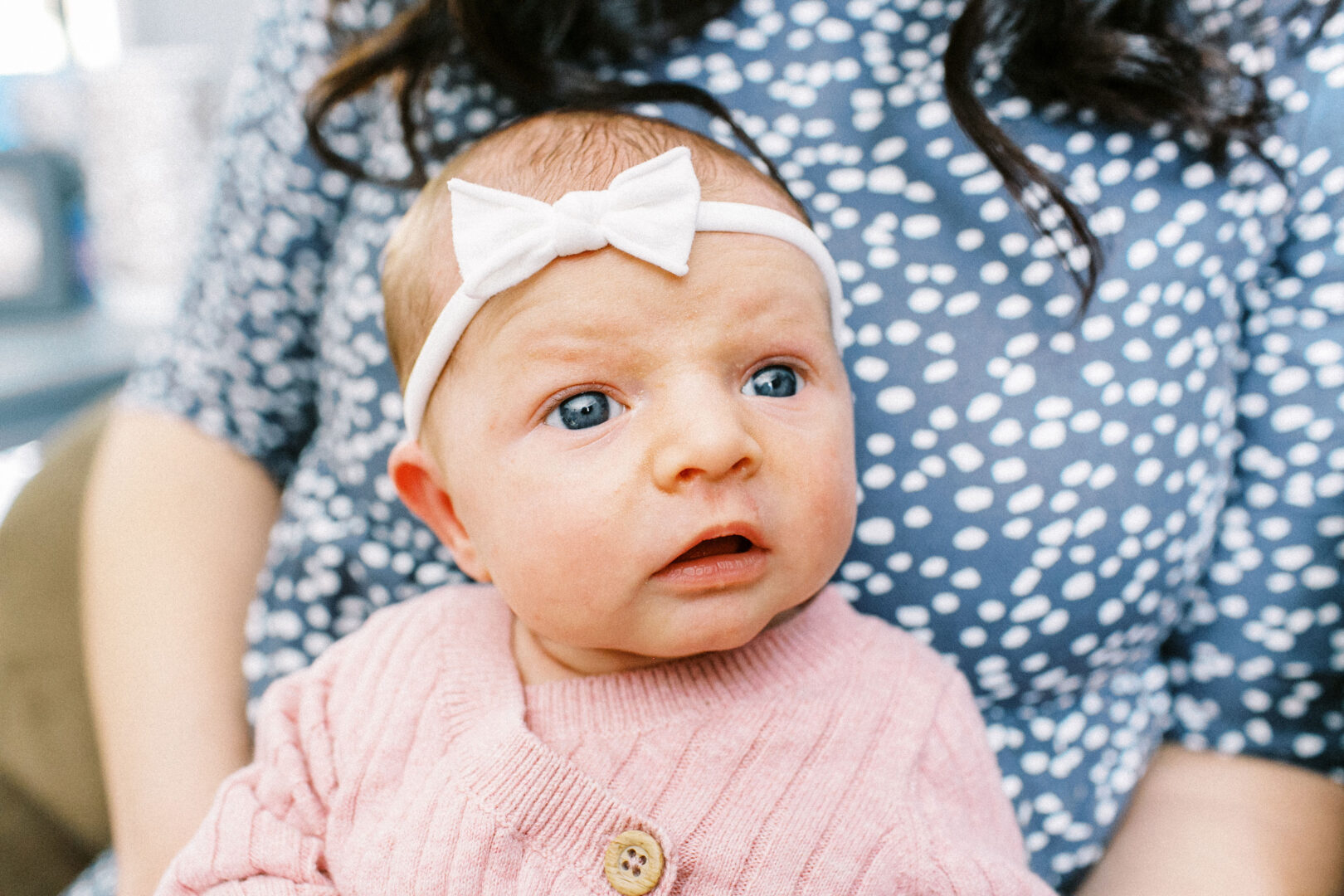 A baby with a white bow headband and pink sweater looks upward while sitting on a person's lap.