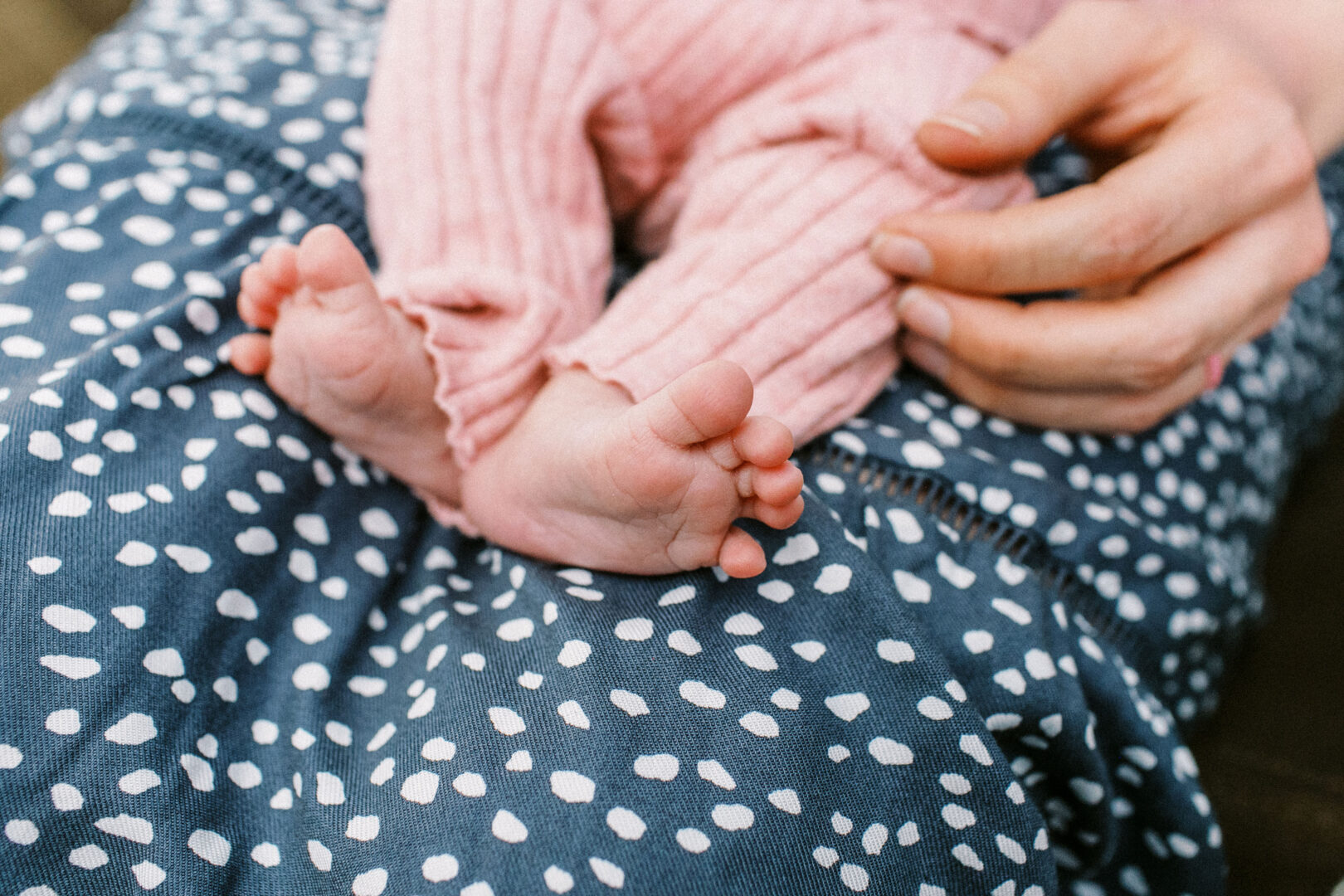 Baby in a pink outfit resting on a person's lap wearing blue patterned fabric, with the person's hand gently placed beside the baby.