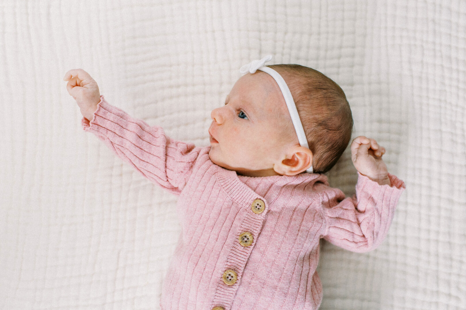 A baby wearing a pink sweater with buttons and a white headband lies on a textured white blanket.