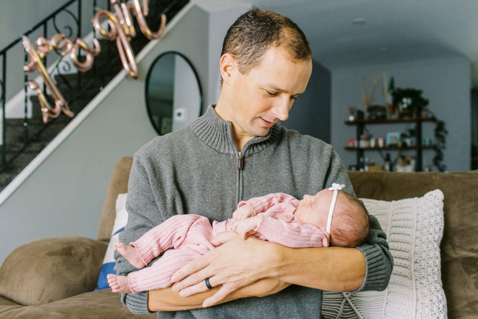 A man holding a baby in pink pajamas in a living room with a staircase in the background.