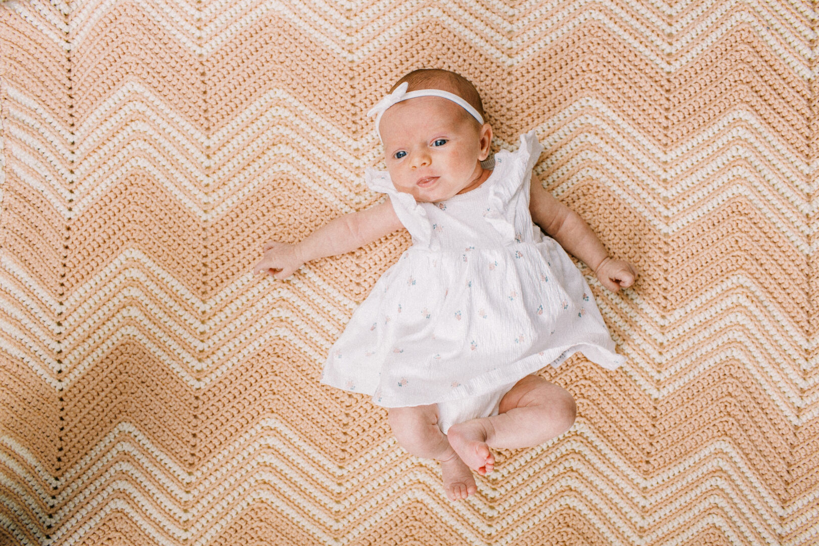 A baby in a white dress and headband lies on a patterned blanket with a zigzag design.