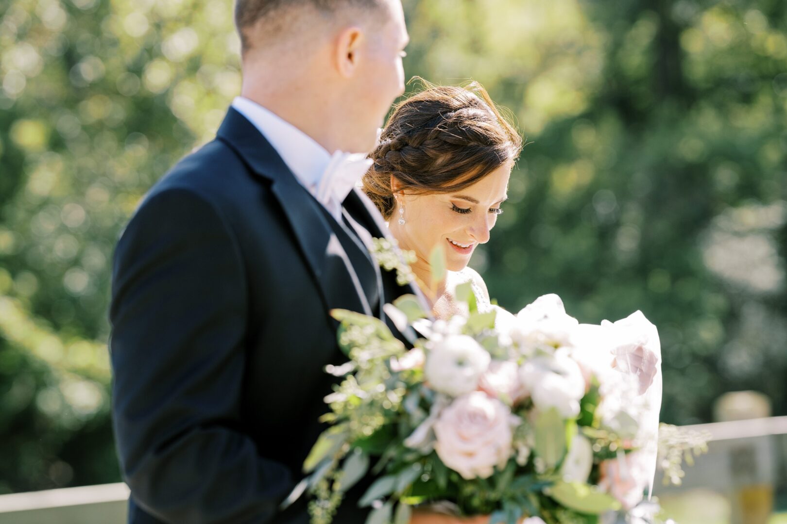 A man in a black suit and a woman in a white dress holding a bouquet of flowers stand outdoors on a sunny day at Barn on Bridge.