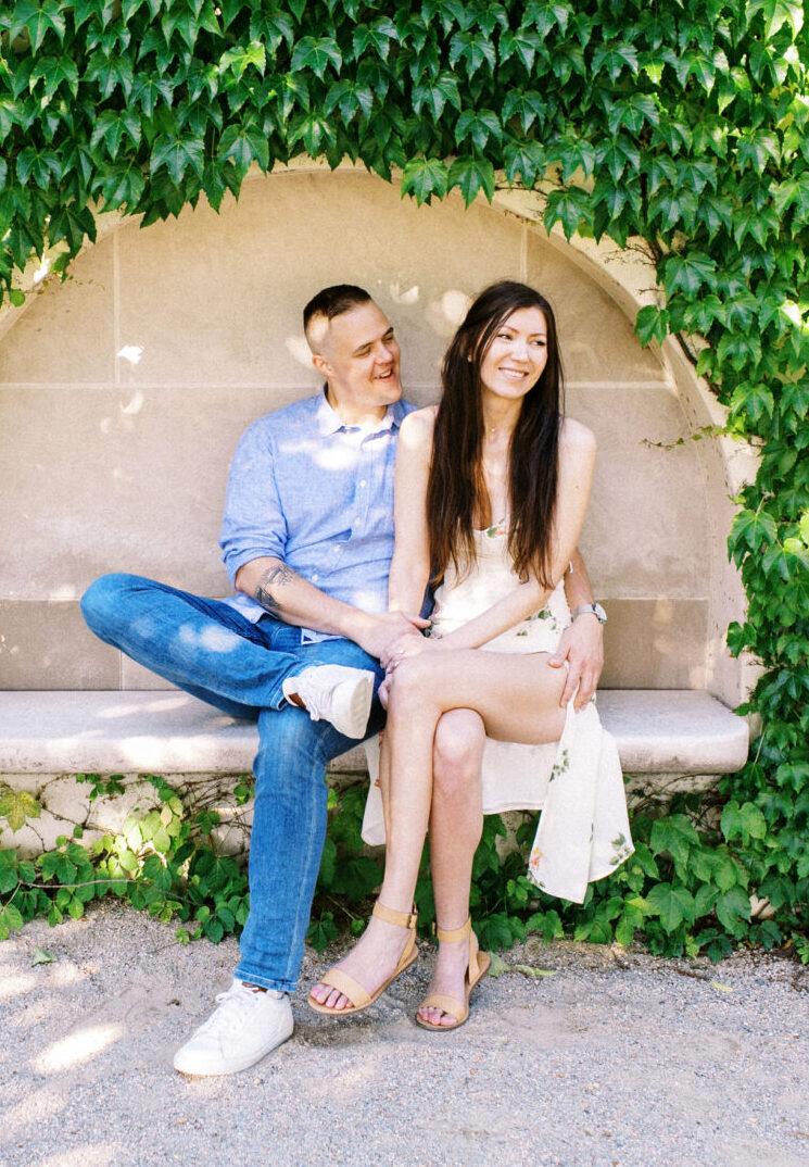 A couple sits on a bench partially covered by climbing plants at Longwood Gardens, smiling and enjoying each other's company on a sunny day—perfect for an engagement session.