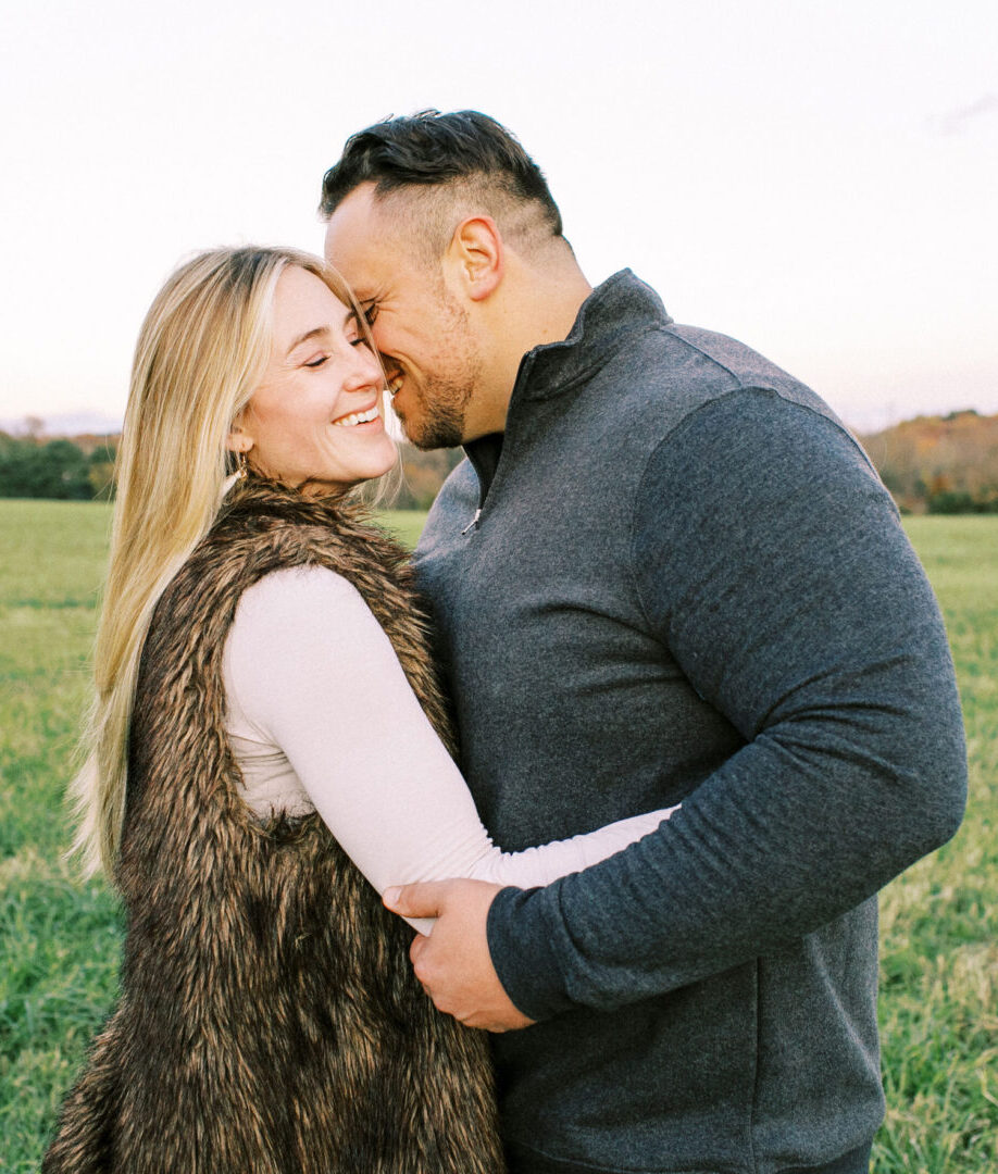 A couple stands in a grassy field, embracing and smiling at each other during their engagement session. The woman wears a fur vest, and the man wears a gray sweater. Trees and a blue sky are in the background, with a charming covered bridge just visible in the distance.