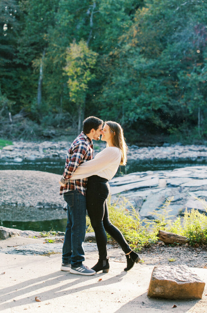 Left: A man and woman stand holding hands, smiling at the camera. Right: During their engagement session, the same couple stands close to each other by a river, sharing an intimate moment in a forested area.