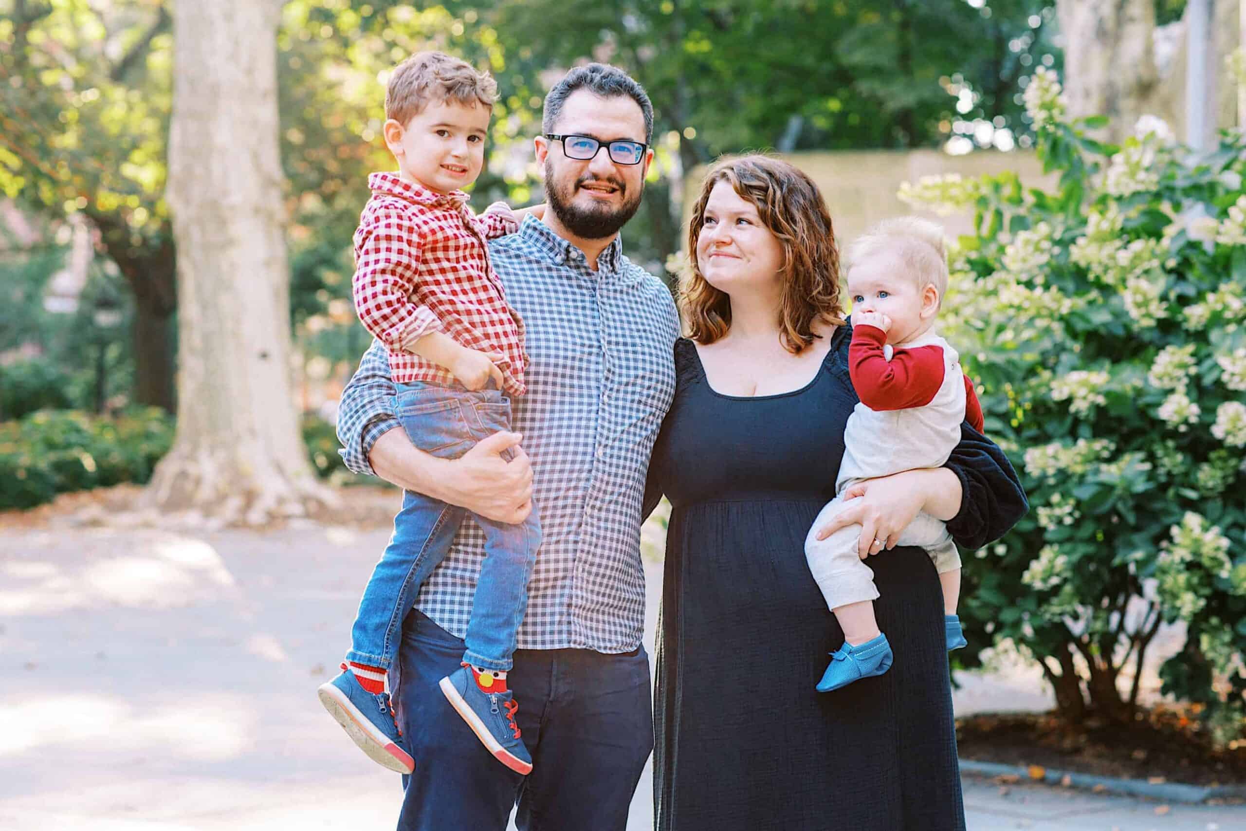 A man and woman stand outdoors with two young children, one in each parent's arms, surrounded by greenery and trees—capturing timeless family photos at Washington Square in Philadelphia.