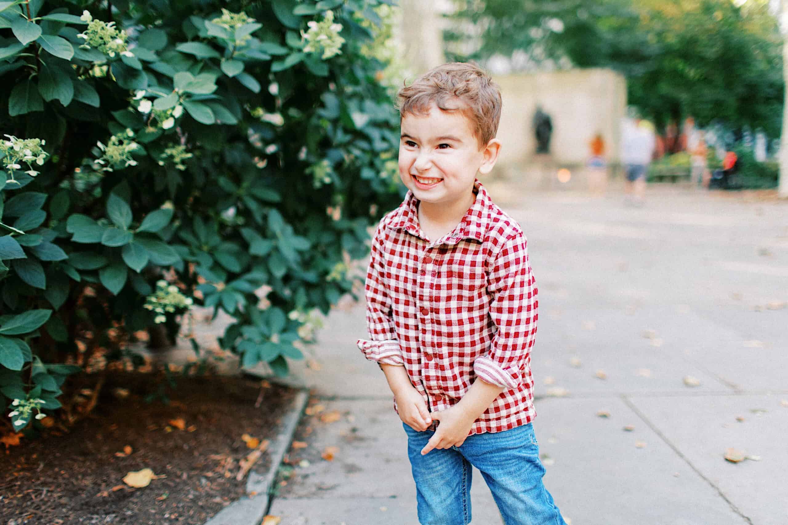 A young child wearing a red plaid shirt and jeans stands on a sidewalk near a bush, smiling and looking to the side—perfect for family photos at Washington Square in Philadelphia.