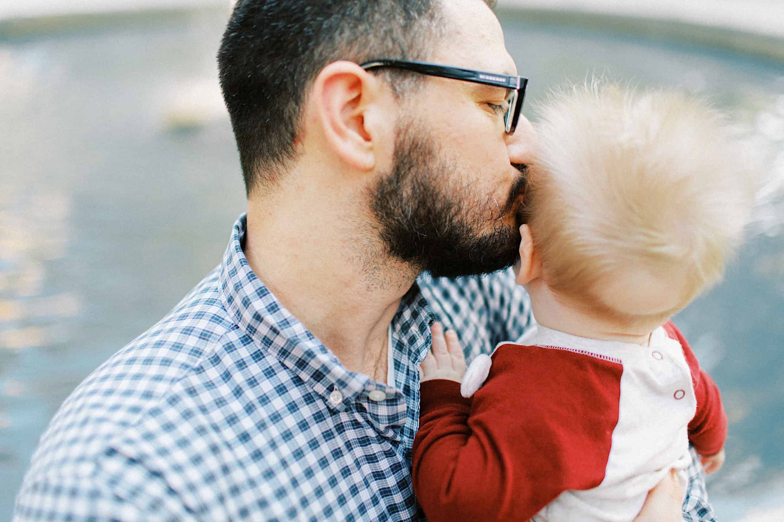 A man with glasses kisses a baby on the head while holding them near a body of water, capturing a tender moment reminiscent of family photos at Washington Square in Philadelphia.
