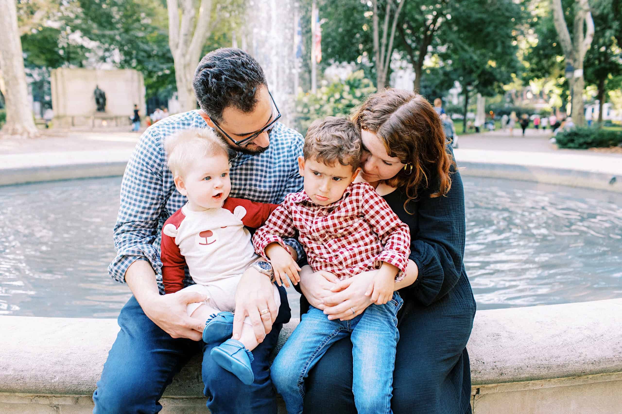 Two adults and two young children sit together on the edge of a fountain for family photos at Washington Square in Philadelphia. One child looks upset while the other is curious. Trees and people fill the background.