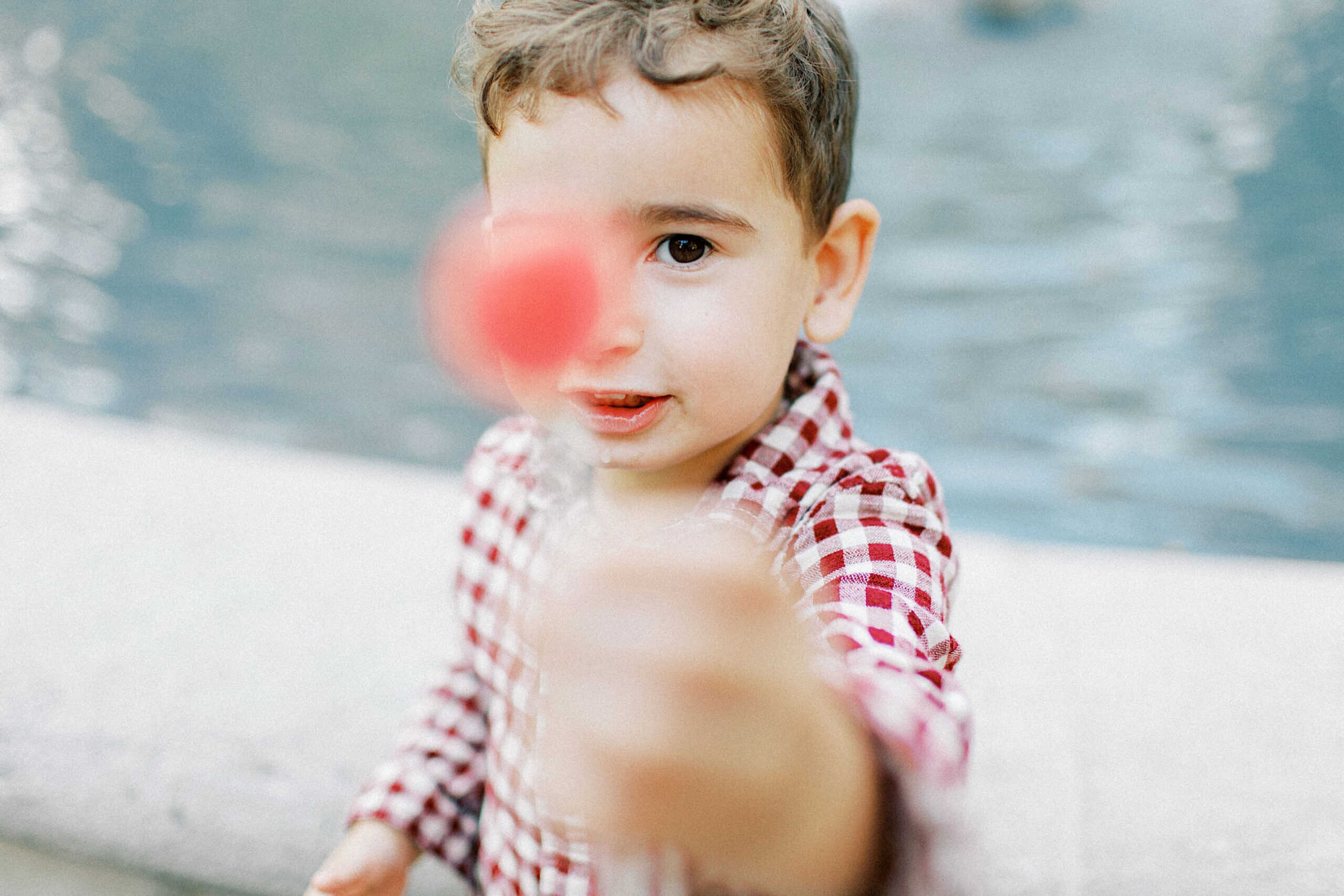 A young boy in a red and white checkered shirt holds a red lollipop toward the camera, capturing the playful spirit of family photos at Washington Square in Philadelphia, as he stands in front of a body of water.
