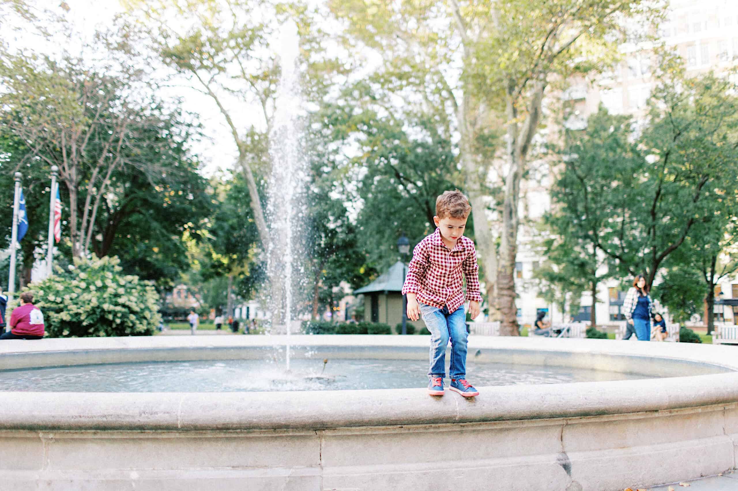 A young boy wearing a red plaid shirt and jeans stands on the edge of a fountain in a park, with water spraying up behind him and trees in the background—capturing classic family photos at Washington Square in Philadelphia.