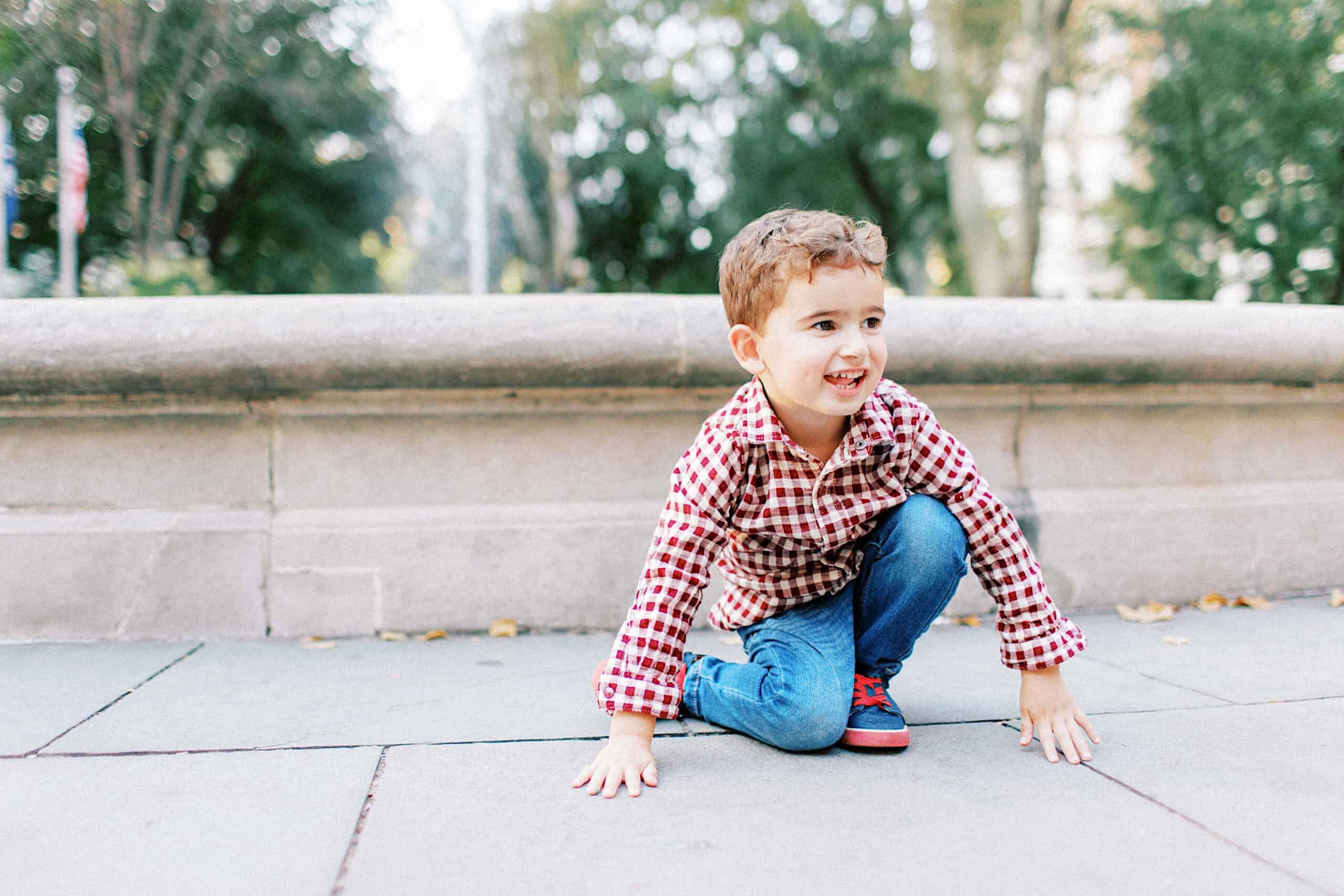 A young boy in a red plaid shirt and jeans squats on a paved area outdoors, smiling, with trees and a stone wall in the background—perfect for family photos at Washington Square in Philadelphia.