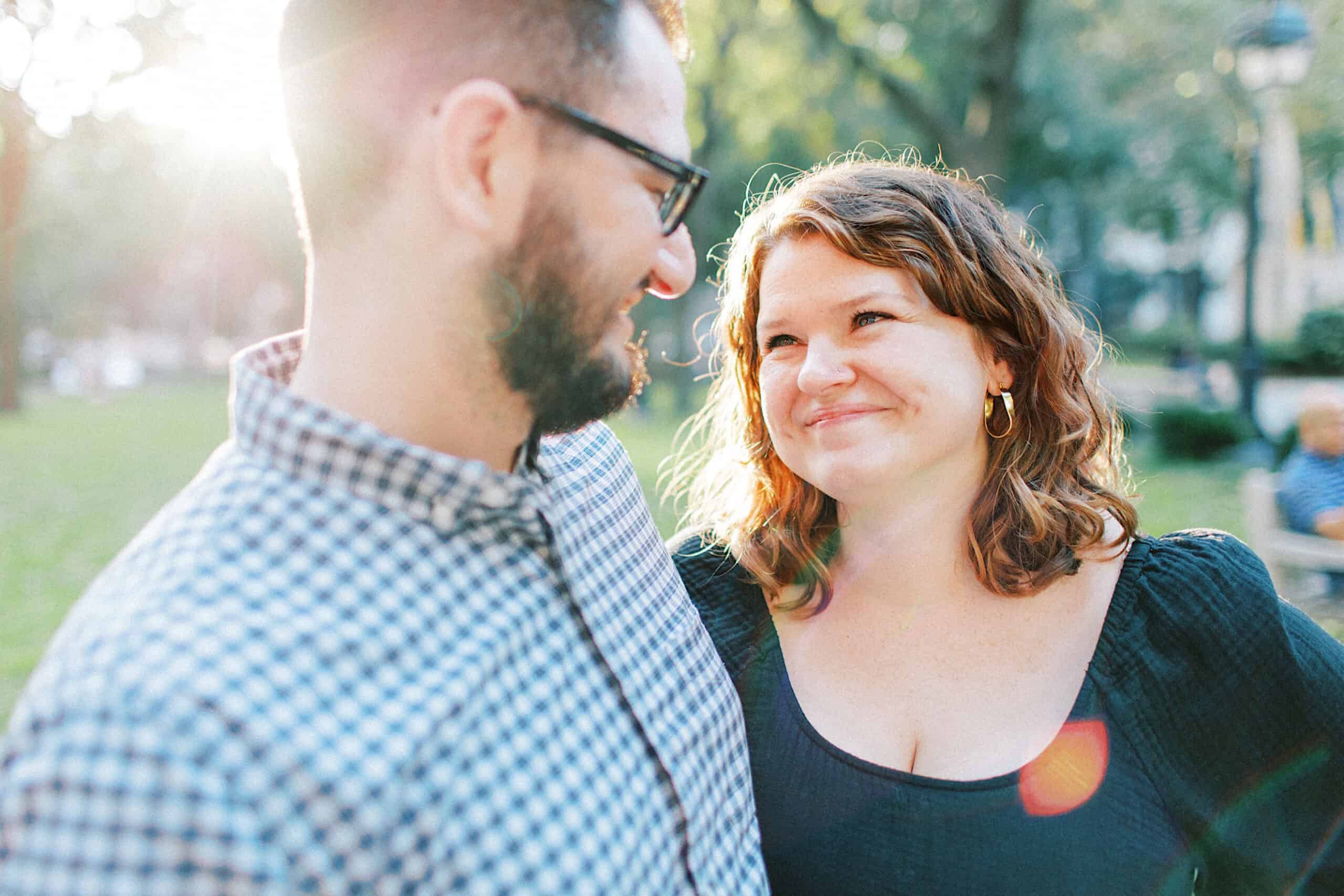 A man and woman stand outdoors in a park, smiling at each other in bright sunlight, perfect for family photos at Washington Square in Philadelphia with trees and people in the background.