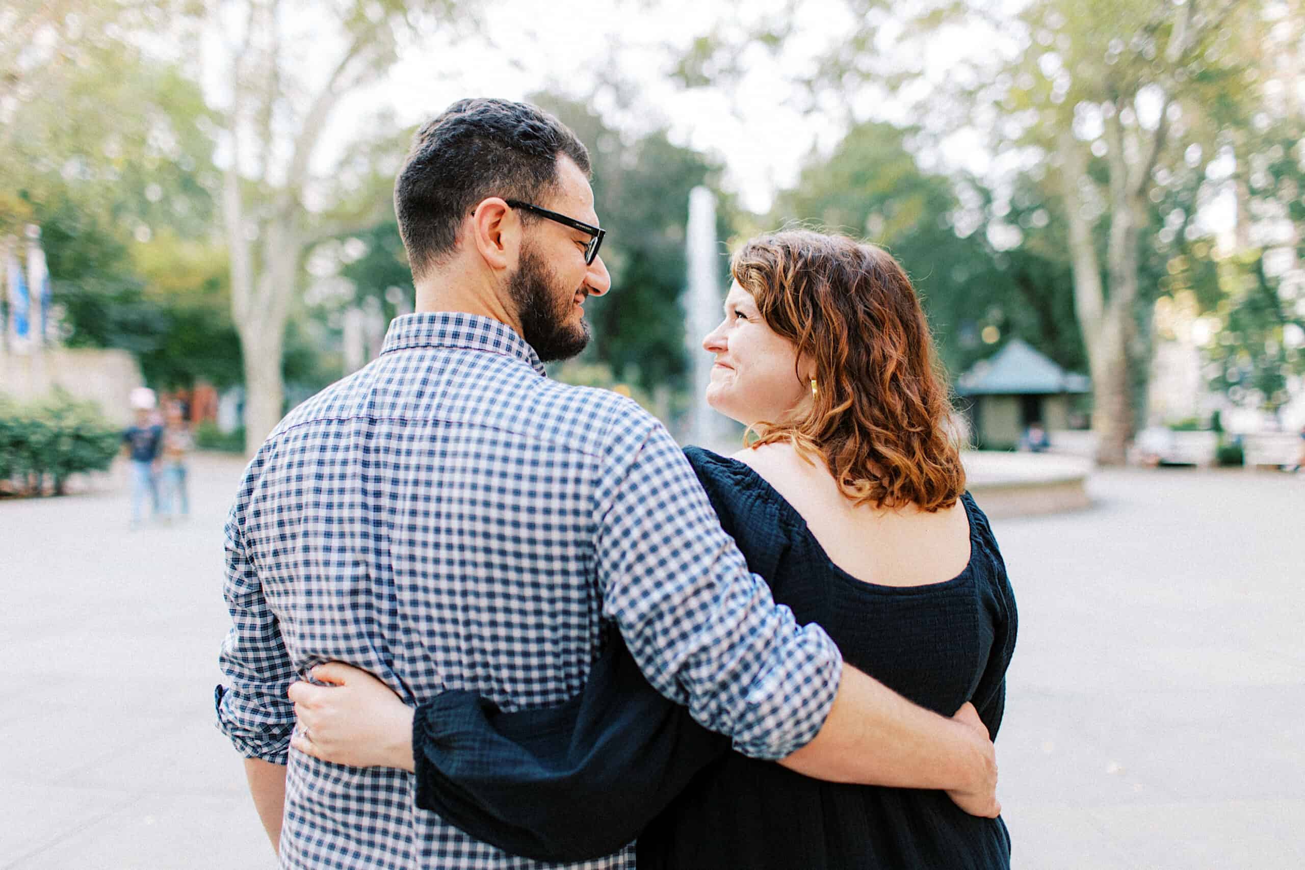 A man and woman stand outside, arm in arm, smiling at each other during family photos at Washington Square in Philadelphia, with trees and a gazebo visible in the background.