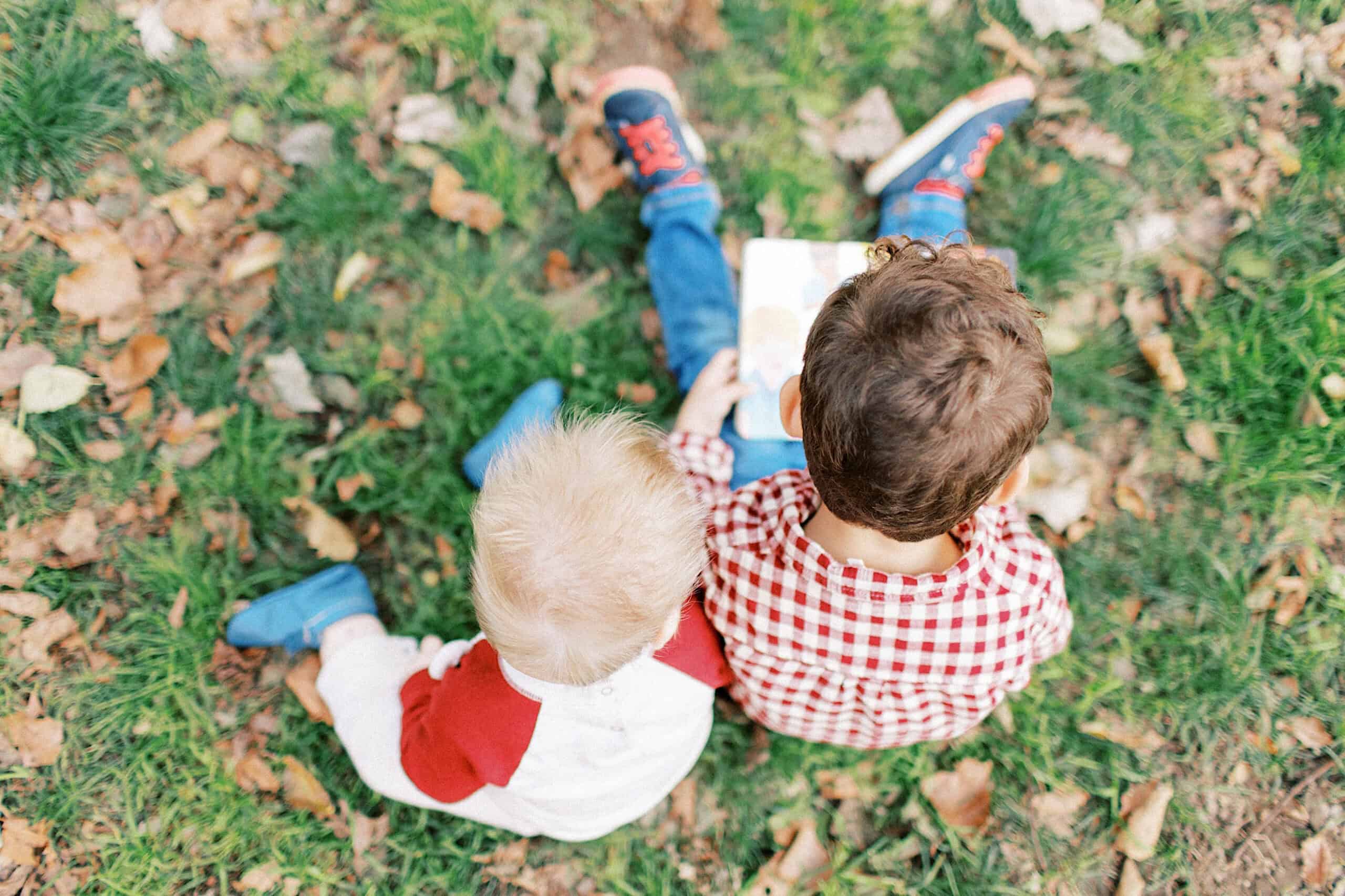 Two young children sit on grass and fallen leaves, seen from above, looking at an open book together—capturing the warmth of family photos at Washington Square in Philadelphia.