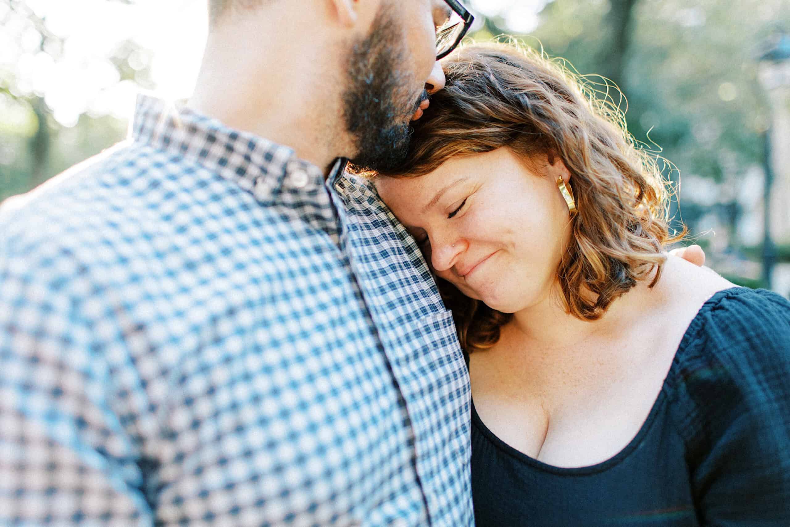 A woman with closed eyes leans contentedly on a man's chest outdoors during family photos at Washington Square in Philadelphia. The man wears glasses and a checkered shirt; the woman wears a black top. Sunlight glows in the background.