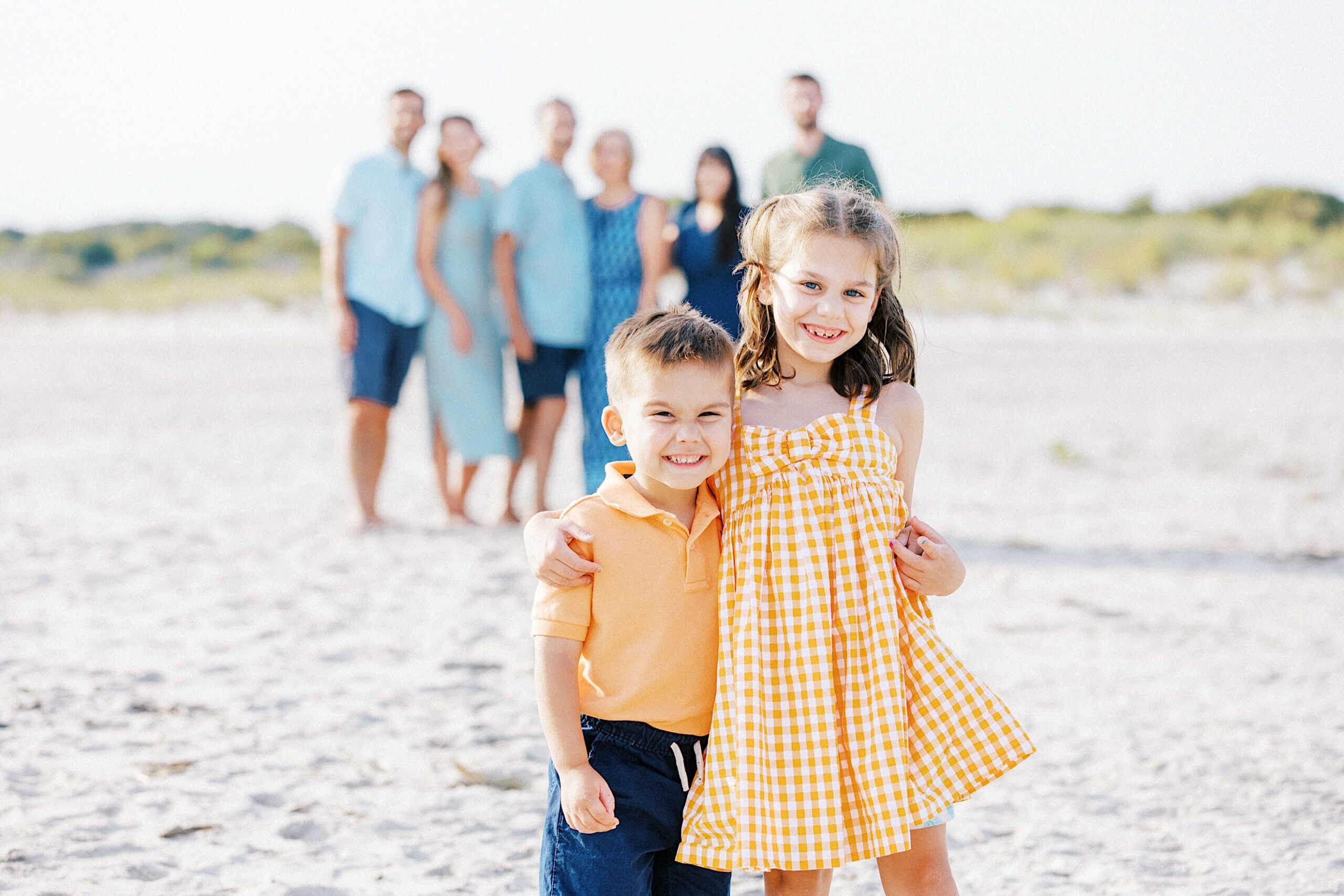 Two children stand smiling on a sandy beach as part of family photos on Cape May Beach in New Jersey, with adults and teens blurred in the background. The sky is clear and the setting is bright.